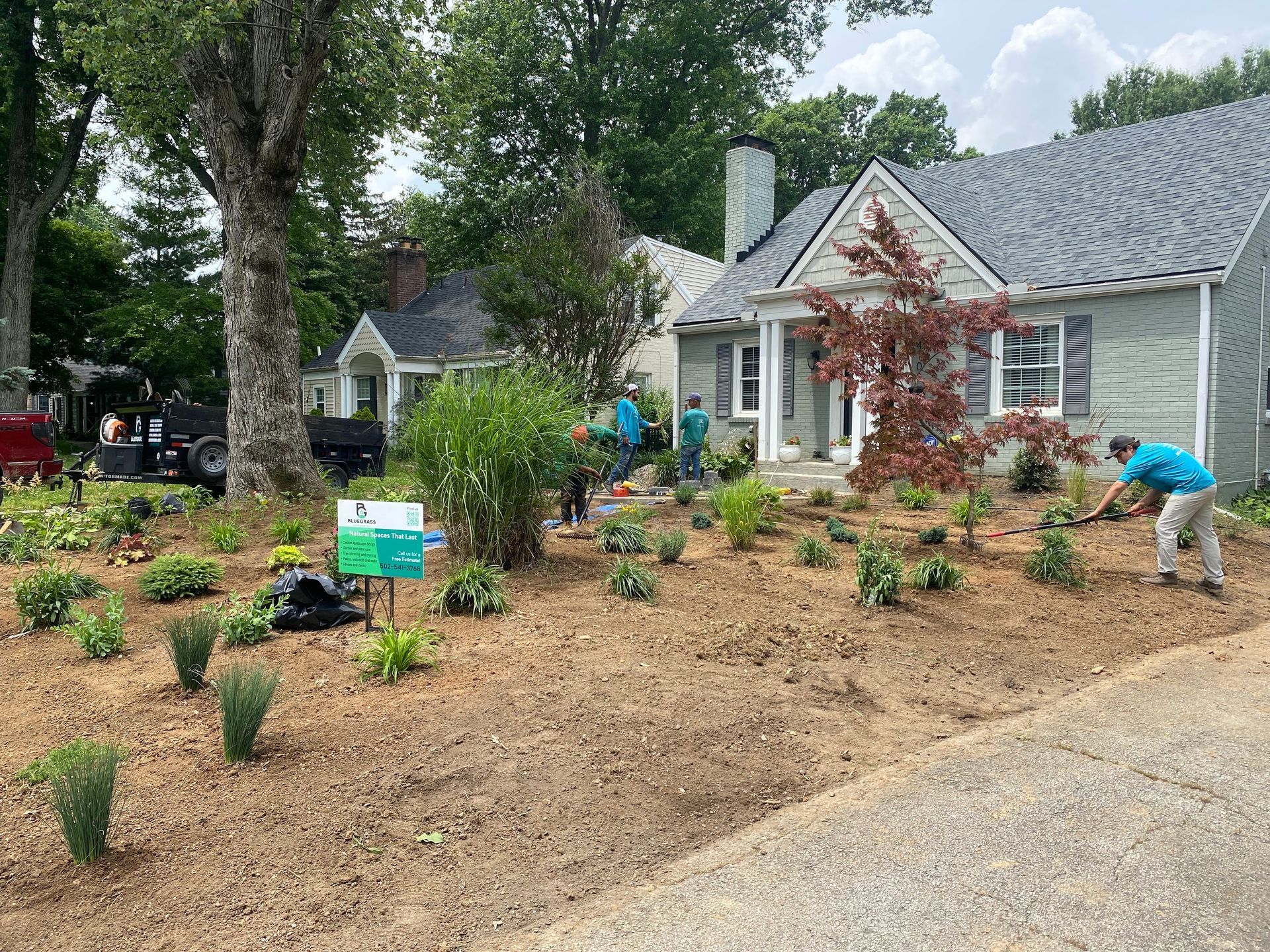 Landscapers planting new greenery in front of a gray house on a sunny day. Mulch and small plants are scattered.