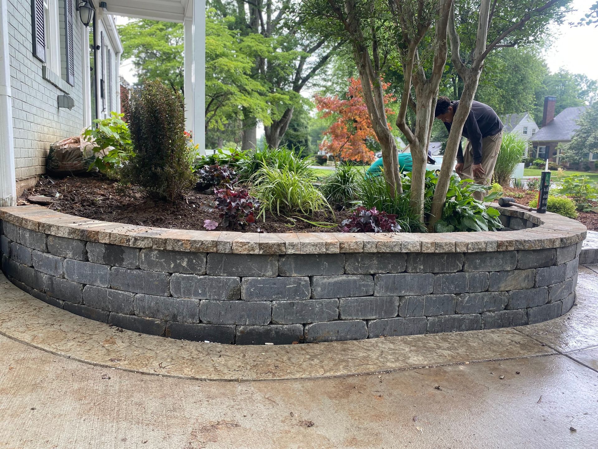 Person gardening in a raised stone planter beside a wet driveway and house