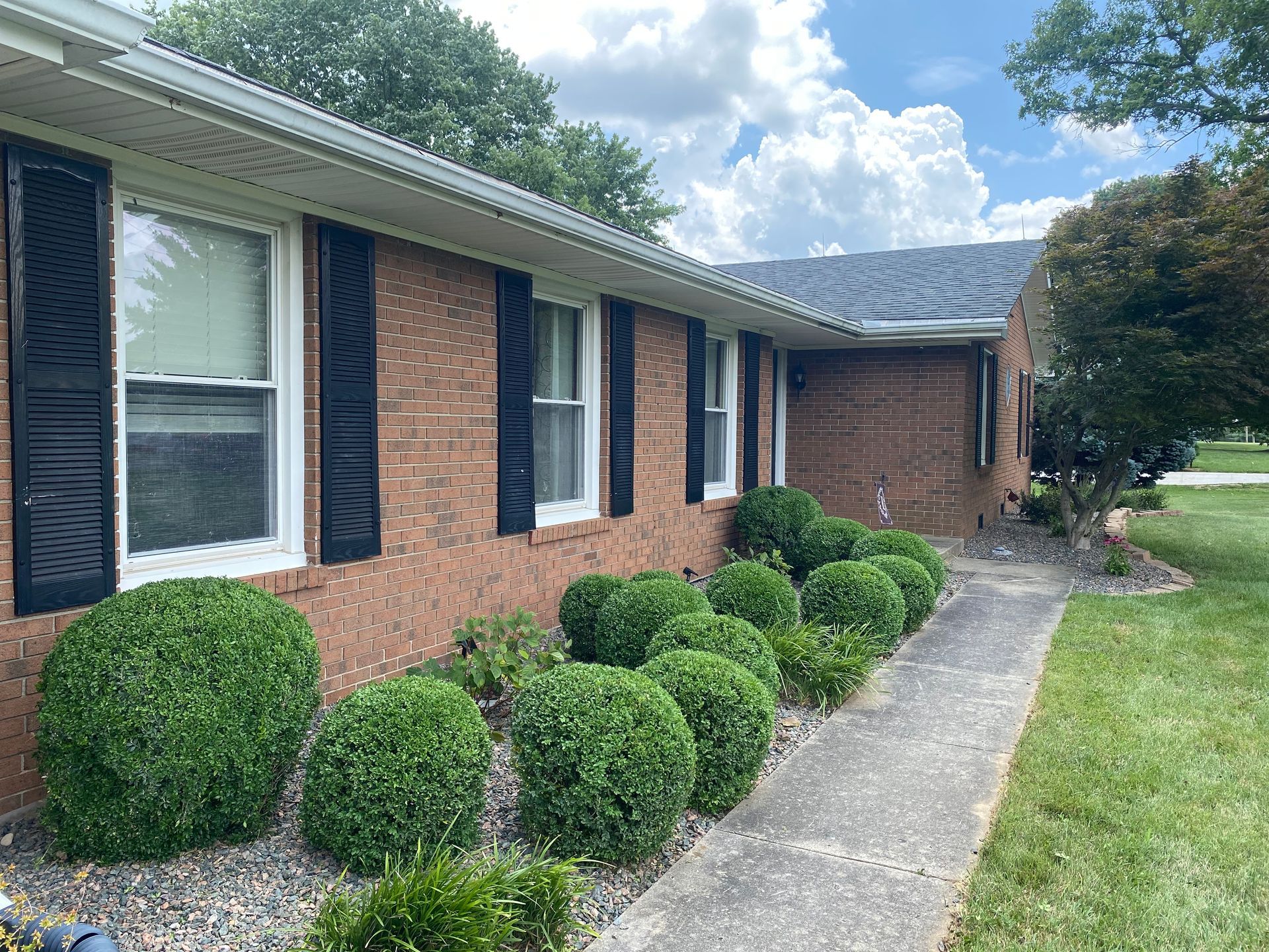 Brick ranch house with black shutters, trimmed bushes, and a concrete walkway beside a green lawn
