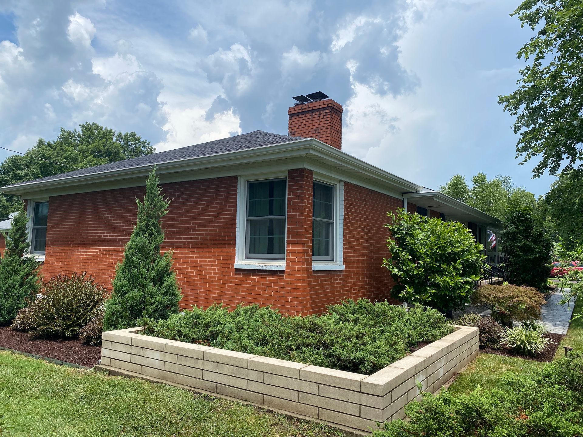 Red brick house with a chimney and white-framed windows. A low brick planter box holds greenery. Overcast sky.