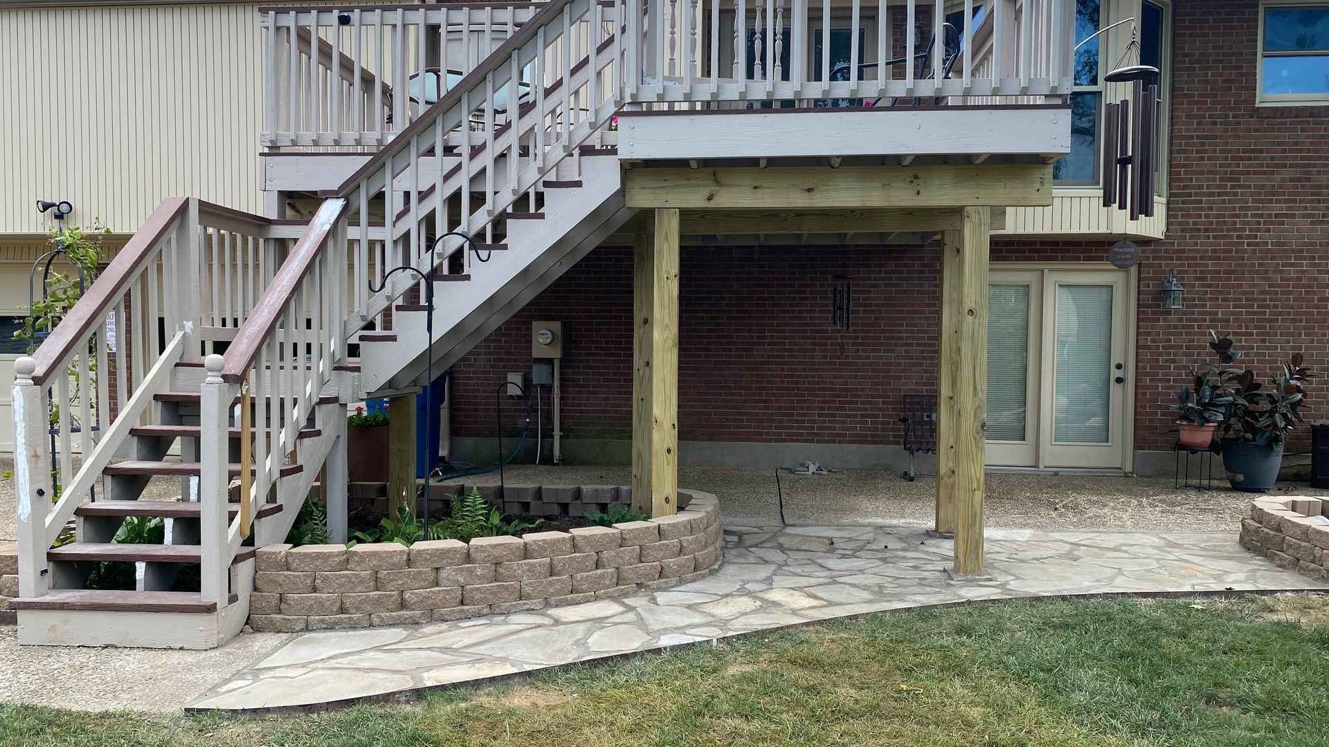 A wooden deck with stairs leading down to a brick patio and a small garden bed in front of a brick house.