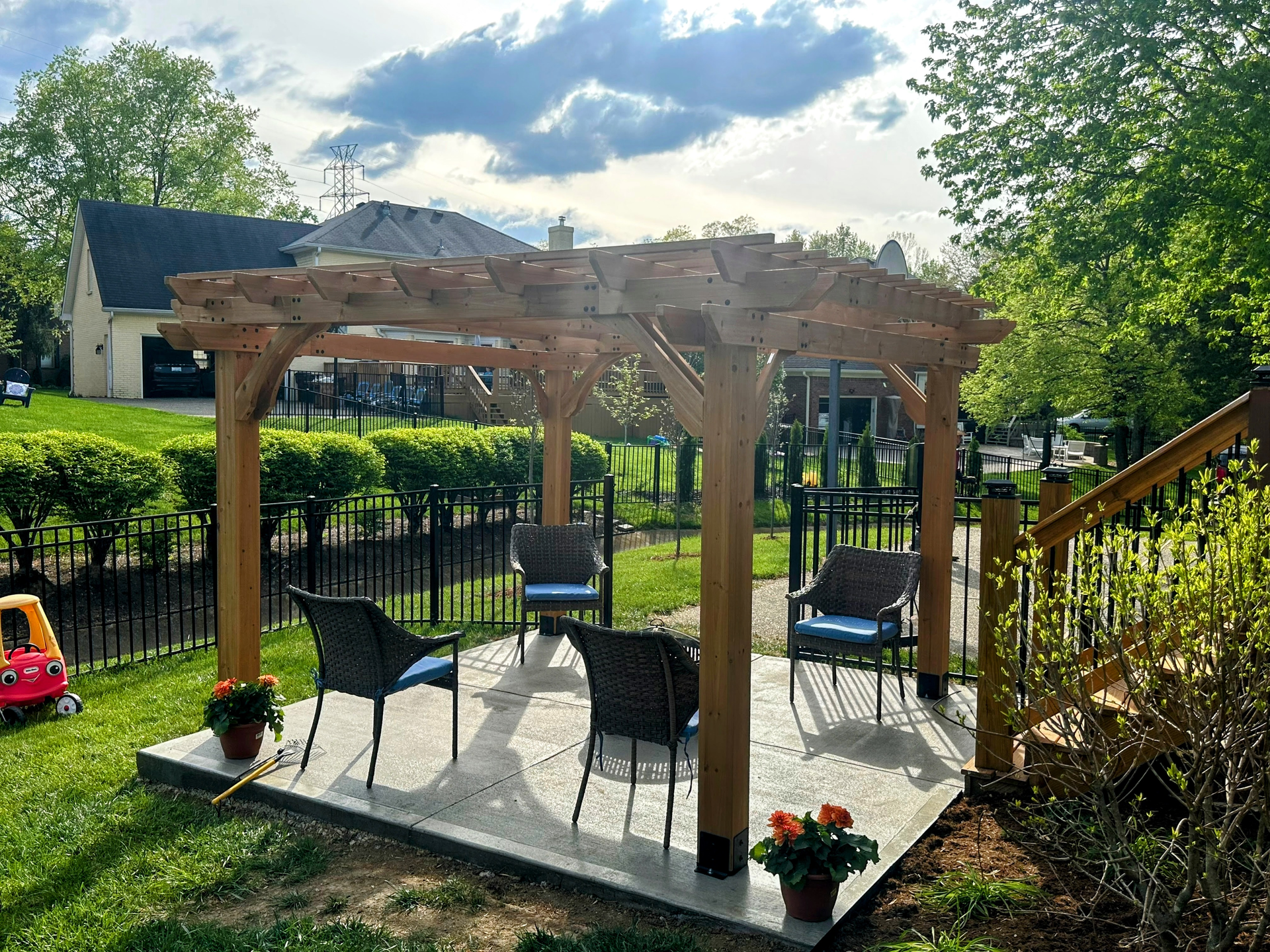 Backyard patio with wooden pergola, chairs, potted flowers, and a fenced lawn in sunlight