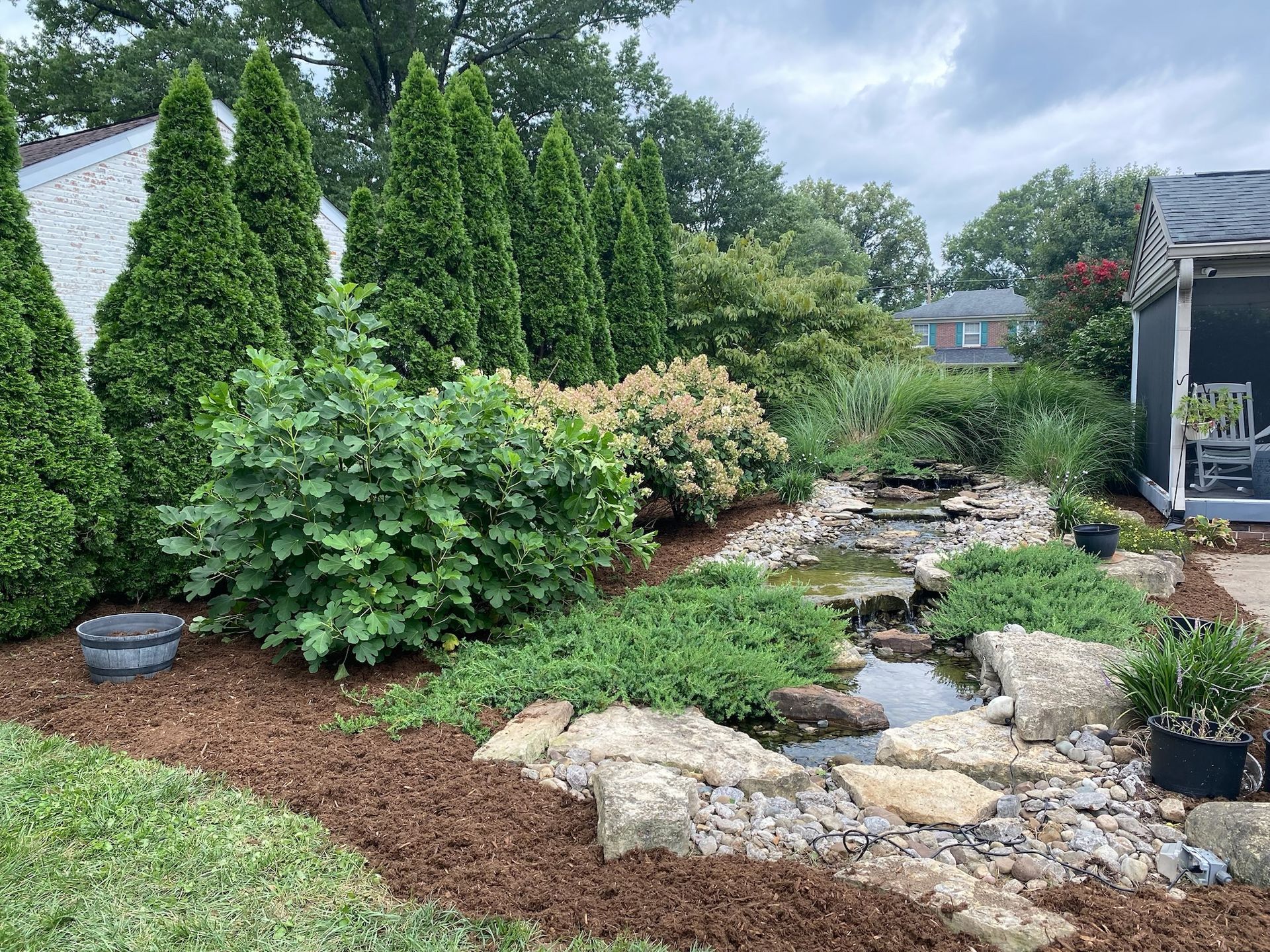 Landscaped garden with stone path, shrubs, tall evergreens, and a small stream beside a house