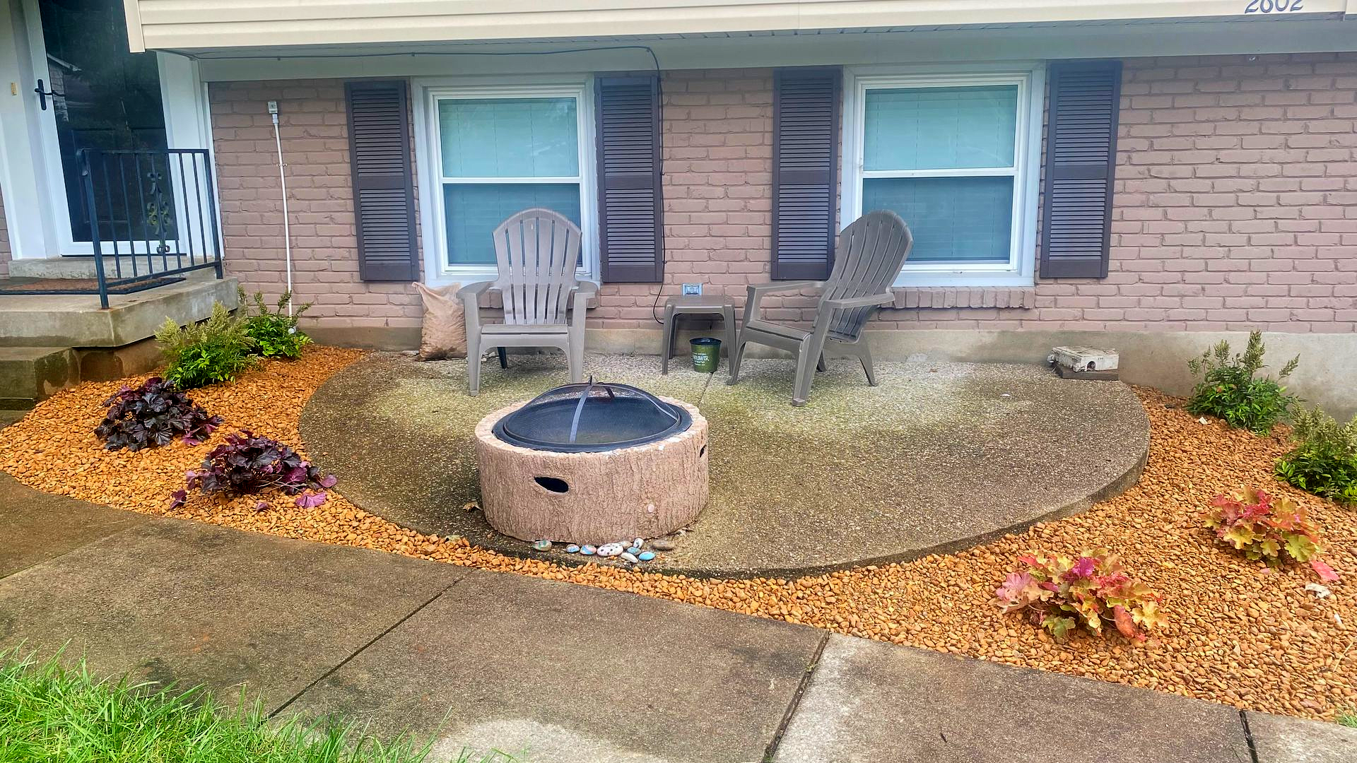 Front yard garden with gravel, mulch, potted chairs, and a circular fire pit in front of a brick house