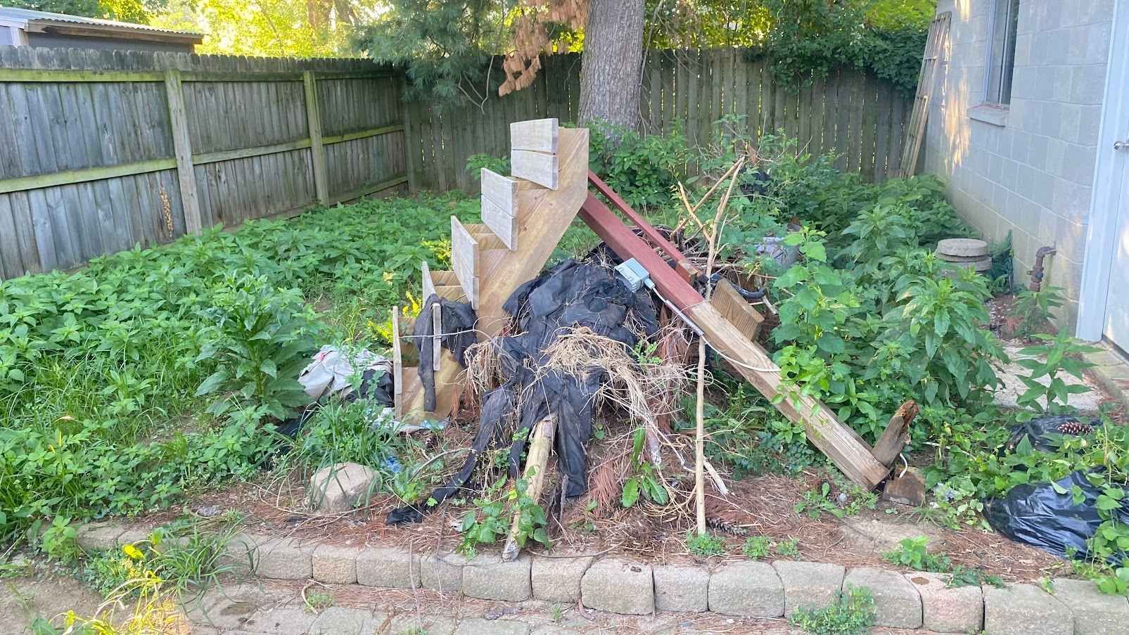 Backyard fire pit with stacked logs and charred wood, surrounded by overgrown greenery and a wooden fence