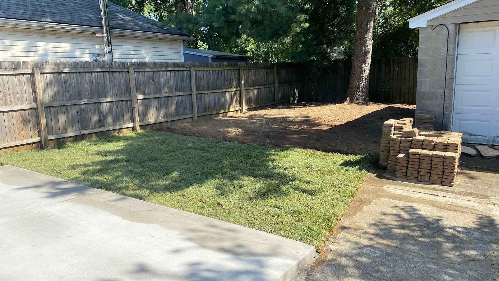 Backyard corner with a wooden fence, patchy grass, driveway, and stacked bricks beside a white shed.