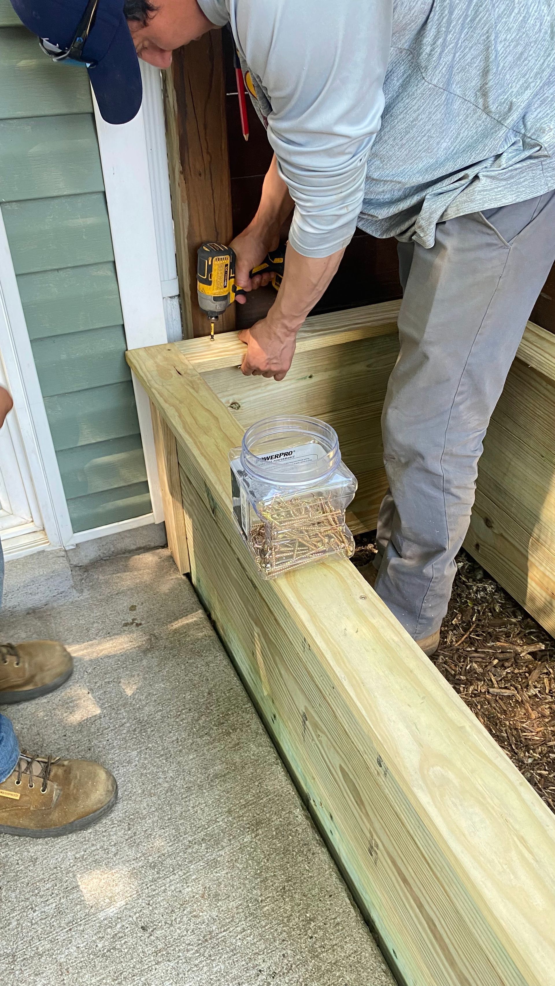 A person in a gray shirt screws into a wooden deck frame. They are working near an entrance.