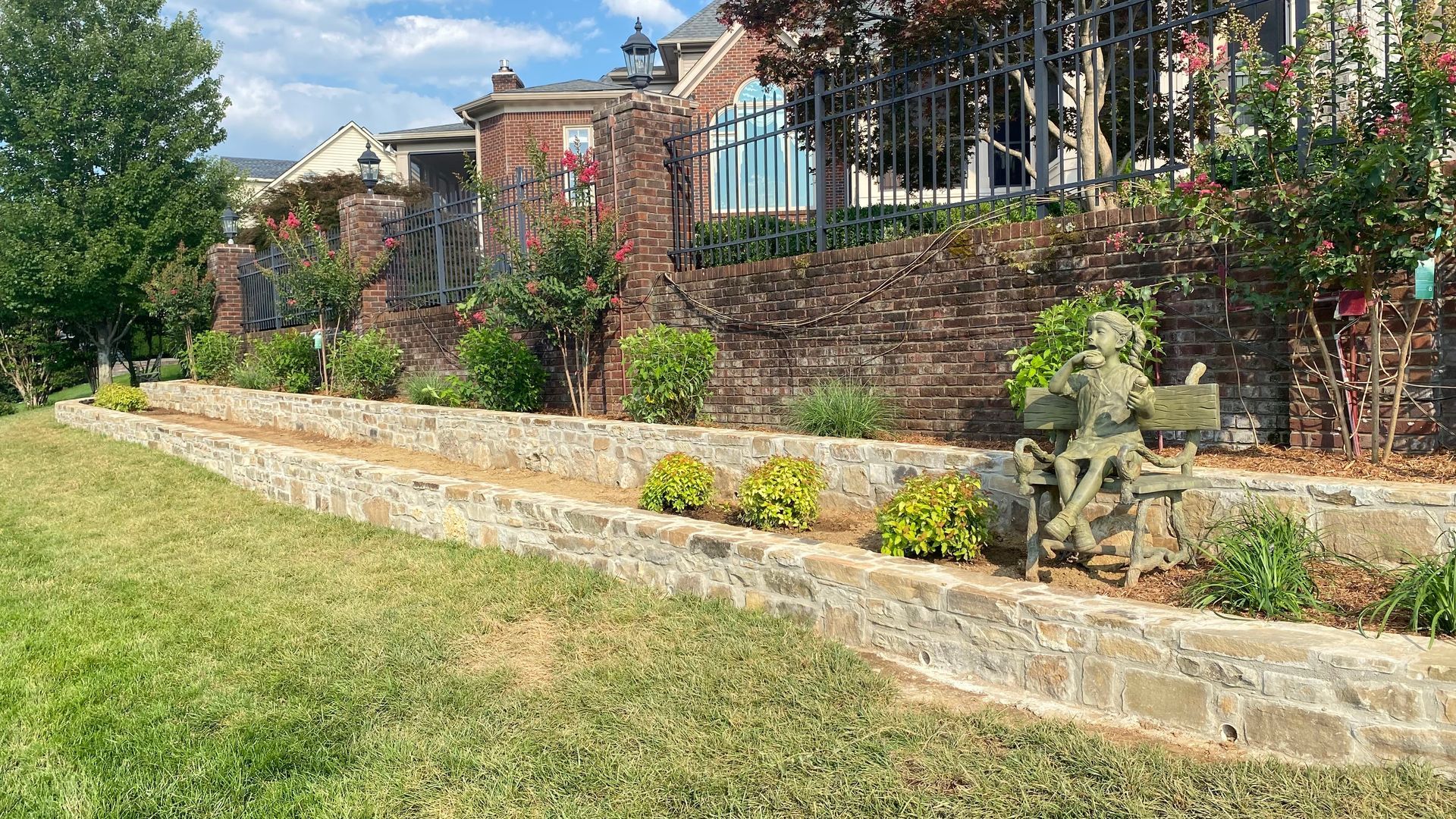 Brick building with stone tiered planters and a bronze statue on a green lawn. The sky is blue.