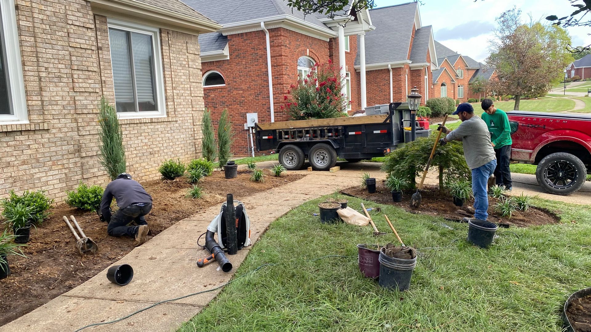Landscapers planting shrubs in front of a brick house. They are using tools like shovels and a blower.
