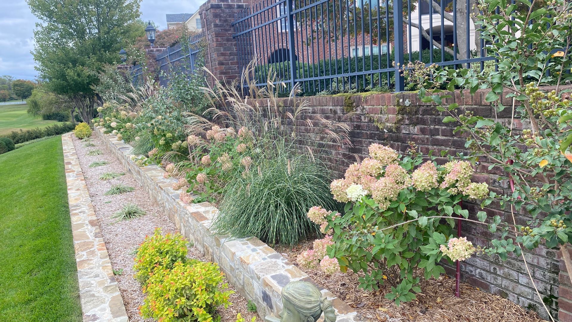 A raised brick flower bed along a wall and fence, filled with tan and green plants. Green grass is visible to the left.