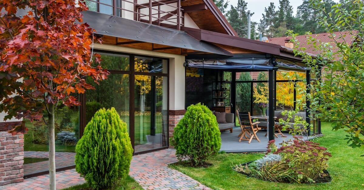 House with large glass windows and a covered patio surrounded by green lawn and trees.