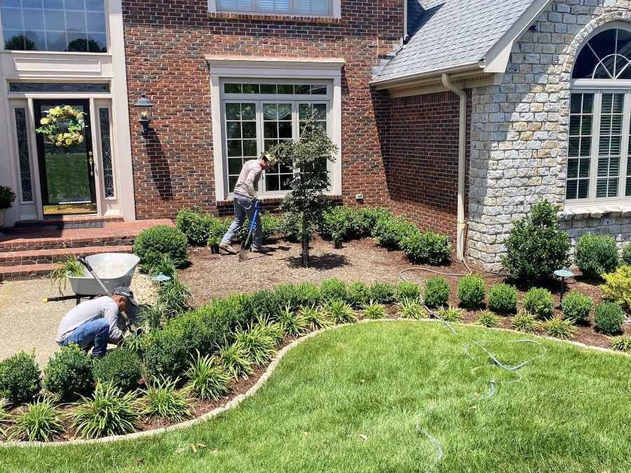 Two people planting a landscaped flower bed beside a brick house and green lawn.