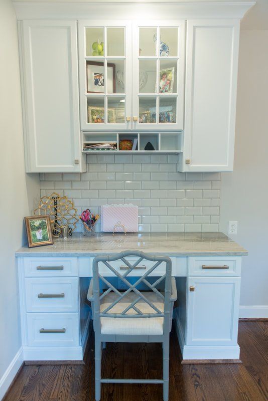 White built-in desk nook with cabinets, glass-front shelves, tiled backsplash, and a chair beneath.