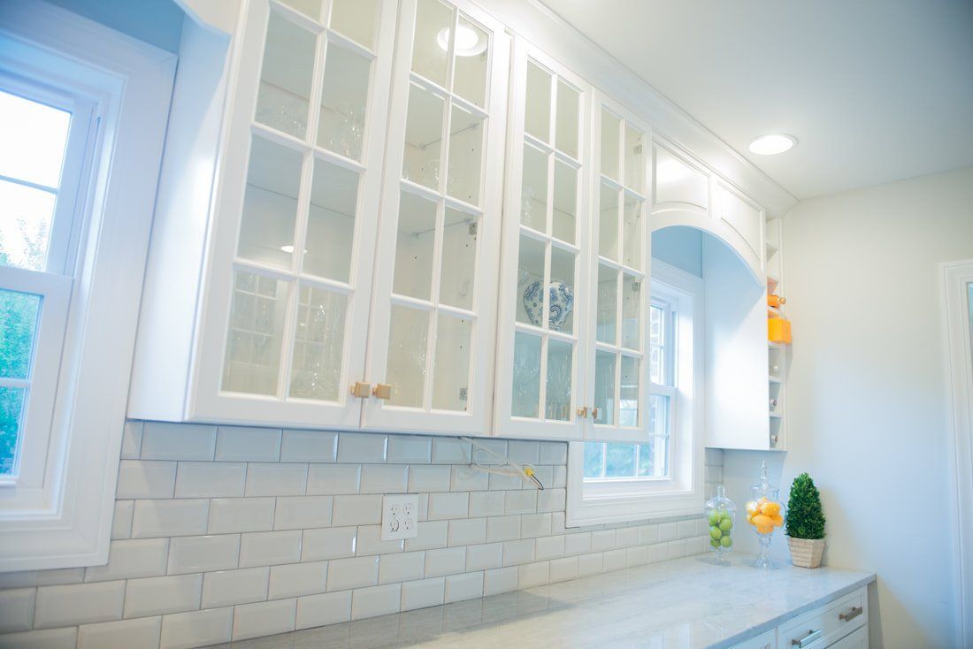 Bright white kitchen with glass-front cabinets, tiled backsplash, and a window over the counter