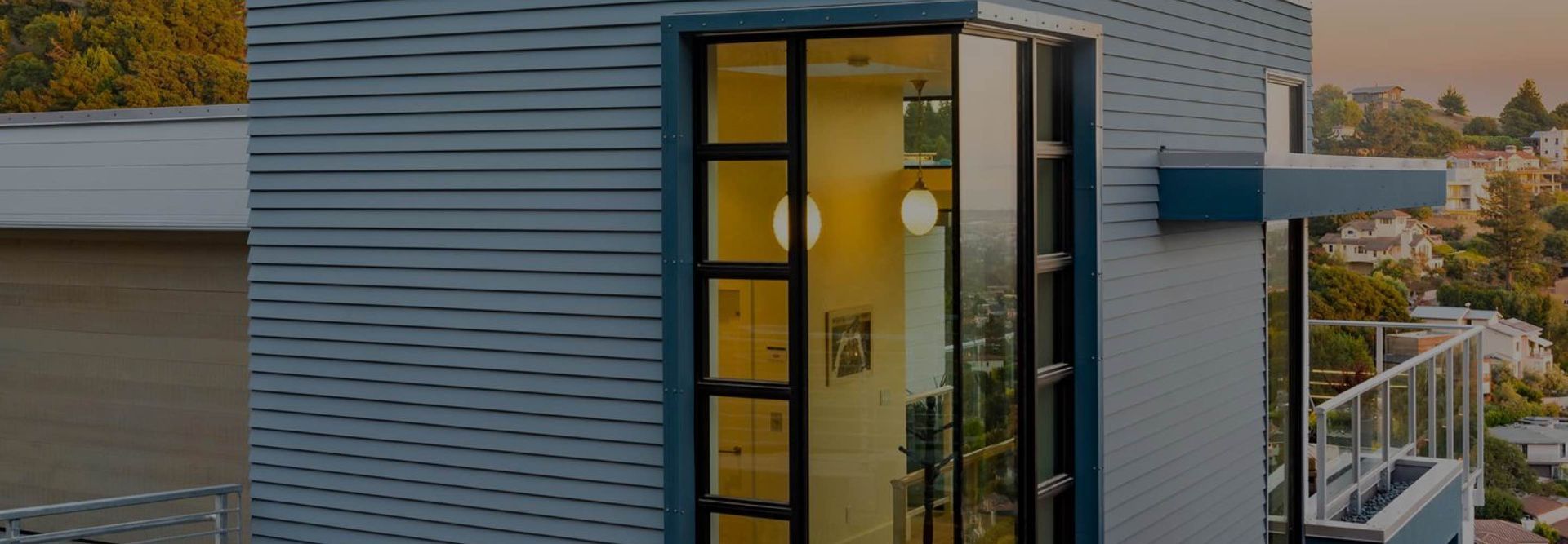 Modern house exterior with blue siding, glass balcony, and warm yellow interior lights