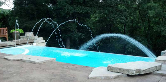 A backyard pool with arcing water jets and stone edging, surrounded by trees.
