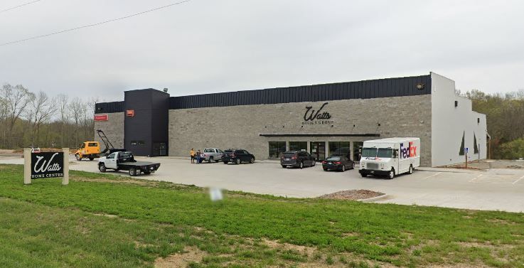Modern industrial building with parked cars, a white van, and a grassy foreground under an overcast sky