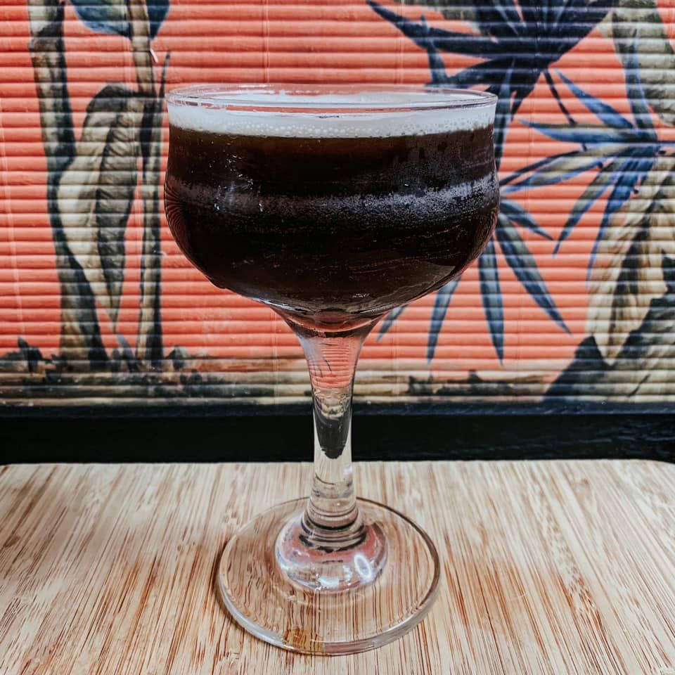 Dark cocktail in stemmed glass, wooden table, tropical wallpaper backdrop.