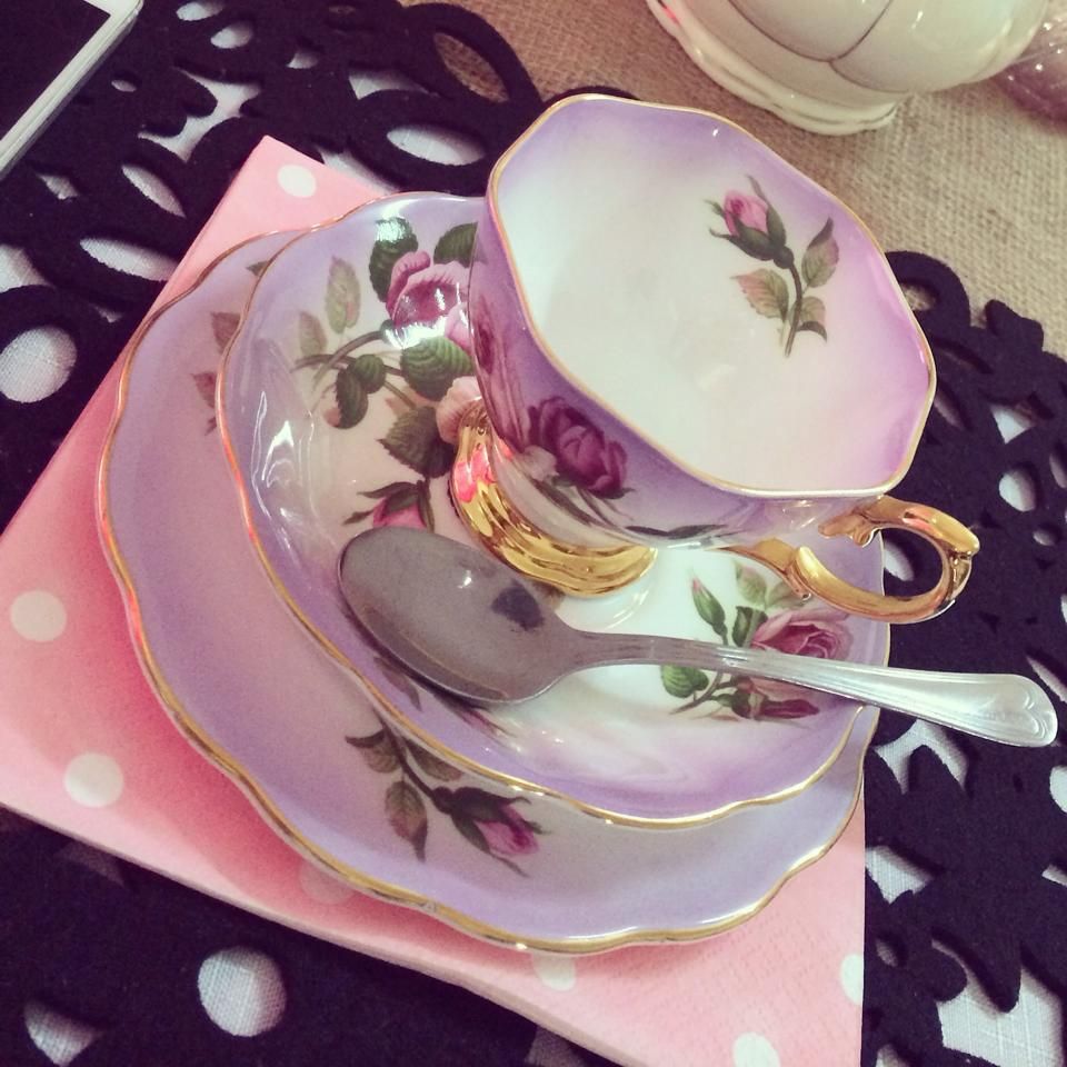 Purple and gold teacup and saucer set with floral design, spoon, on pink napkin, black patterned surface.