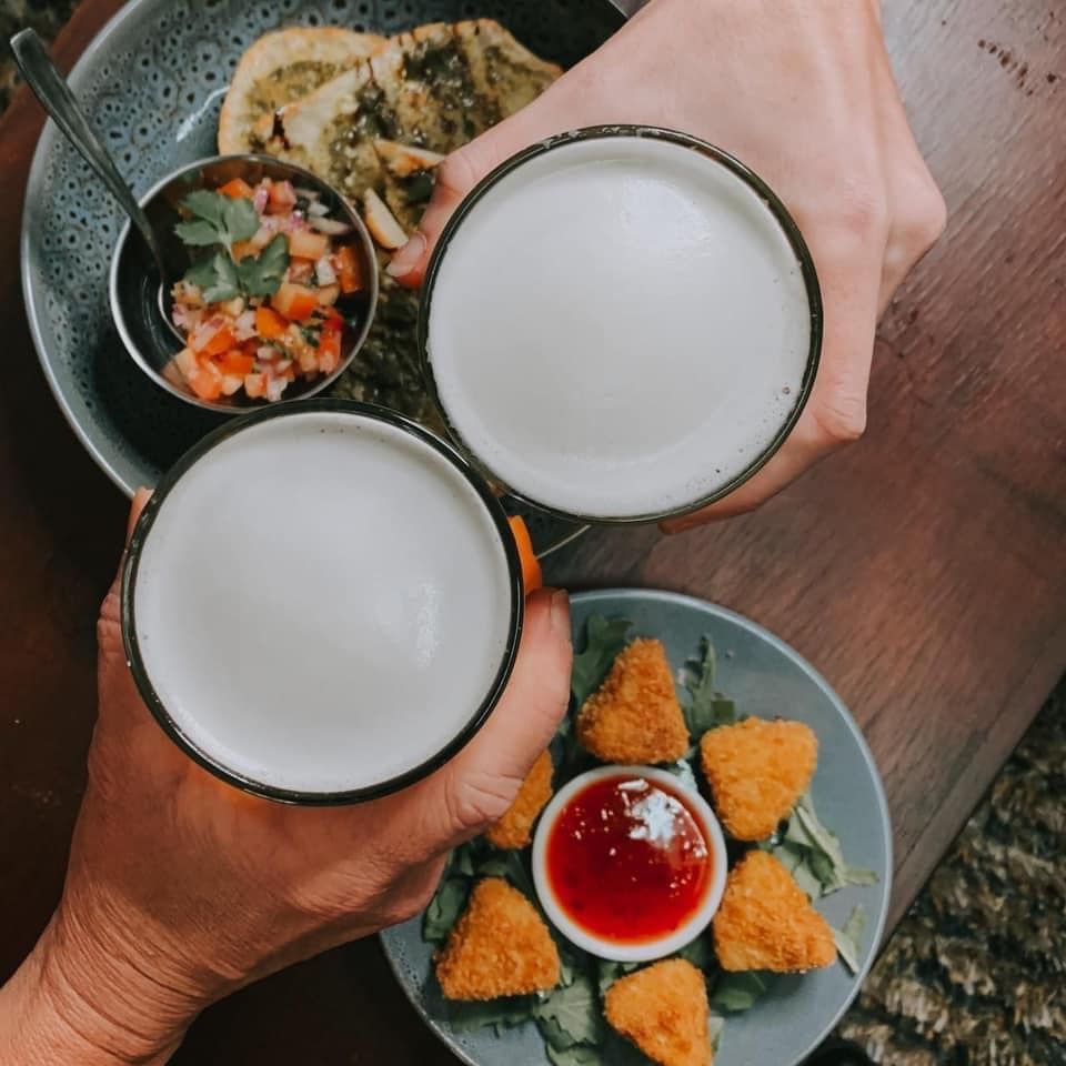 Two people clink beer mugs above plates of food: fried cheese and a dip, and pesto-covered flatbread with salsa.