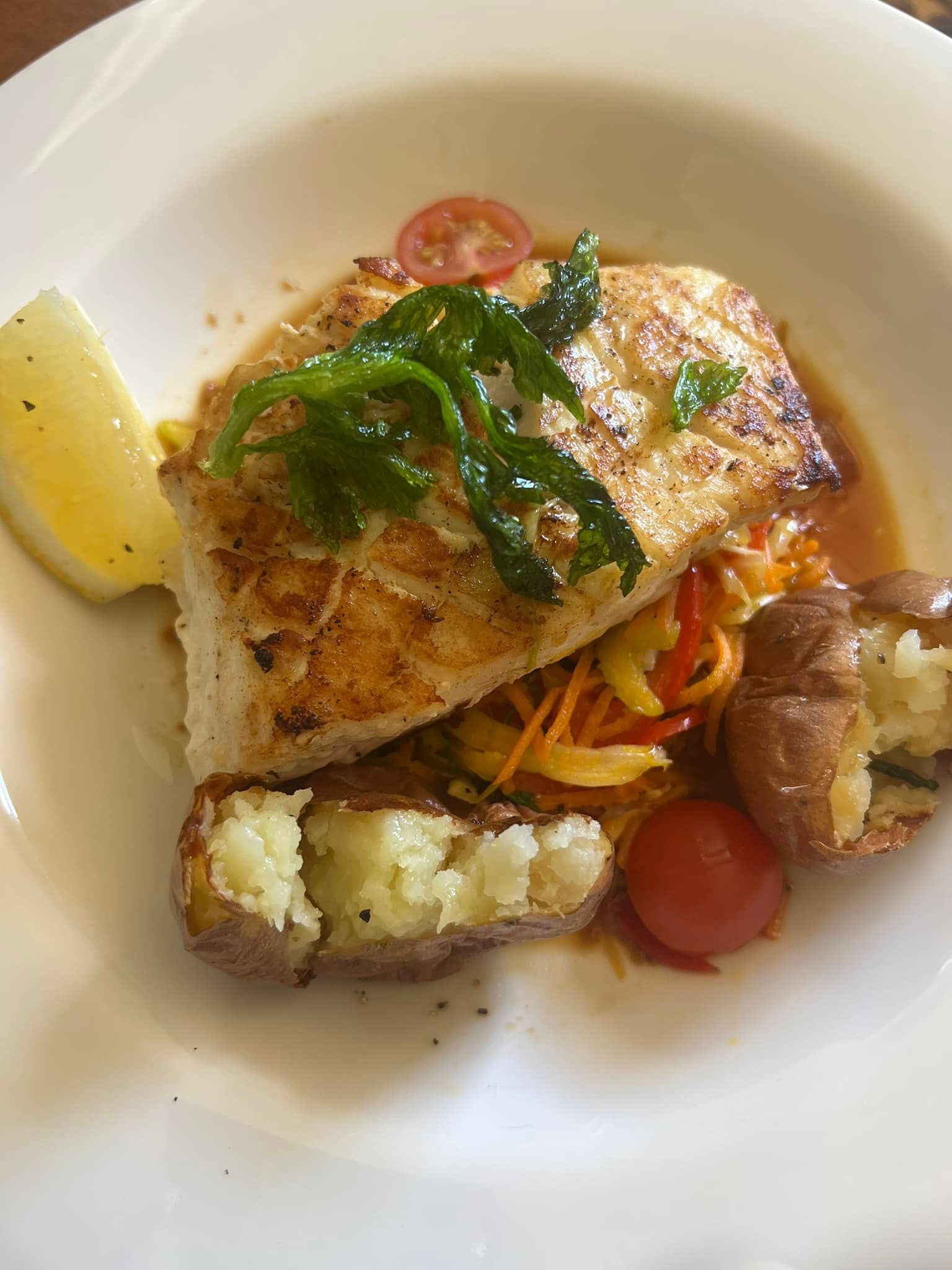 Seared fish fillet with roasted potatoes, vegetables, herbs, tomato, and lemon wedge on a white plate.