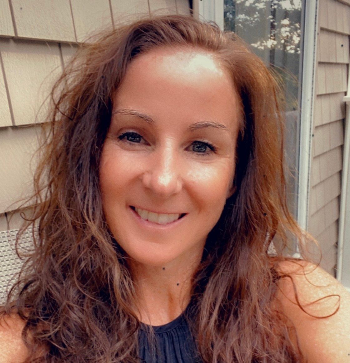A woman with long brown hair is smiling in front of a house.