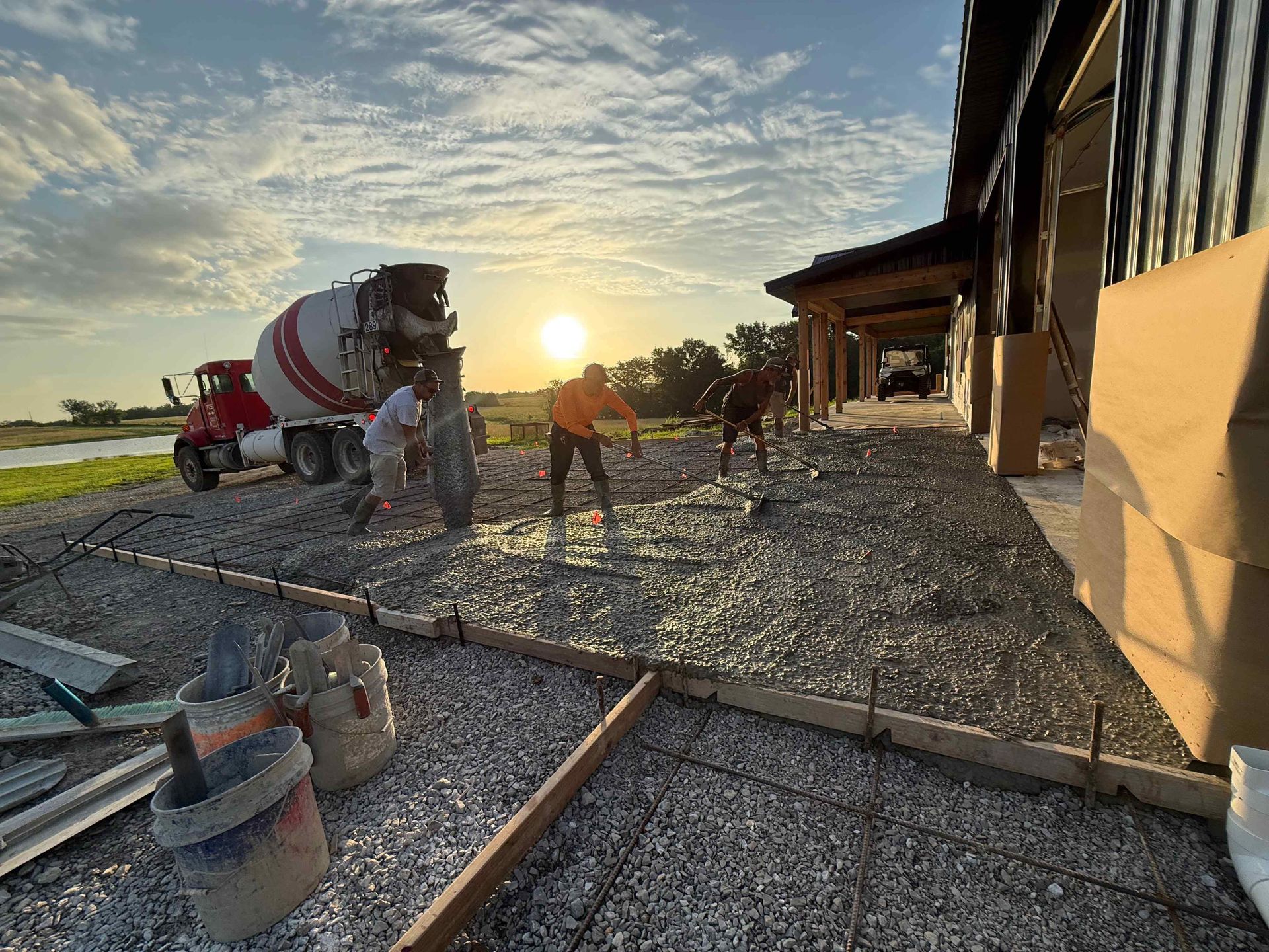 Workers pouring concrete at sunset beside a building on a gravel site