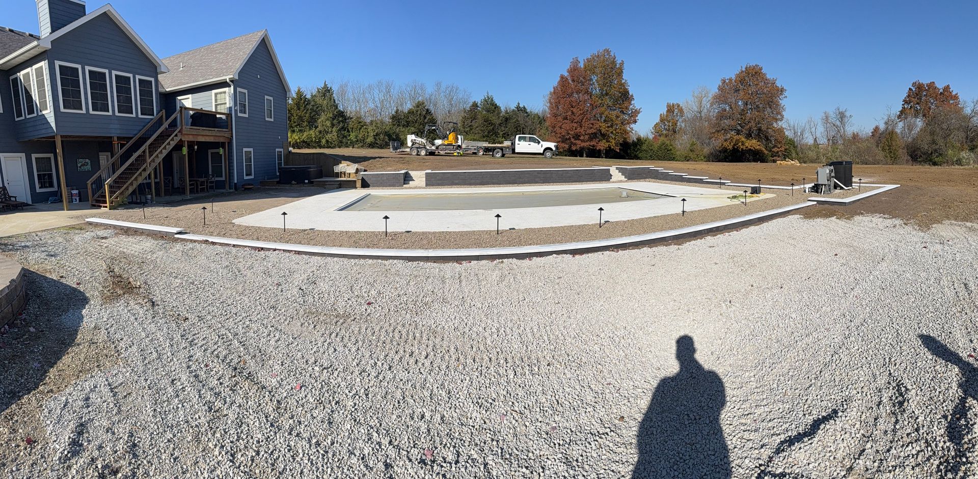 Gravel driveway under construction beside a gray house, with curved concrete edging and autumn trees in back.