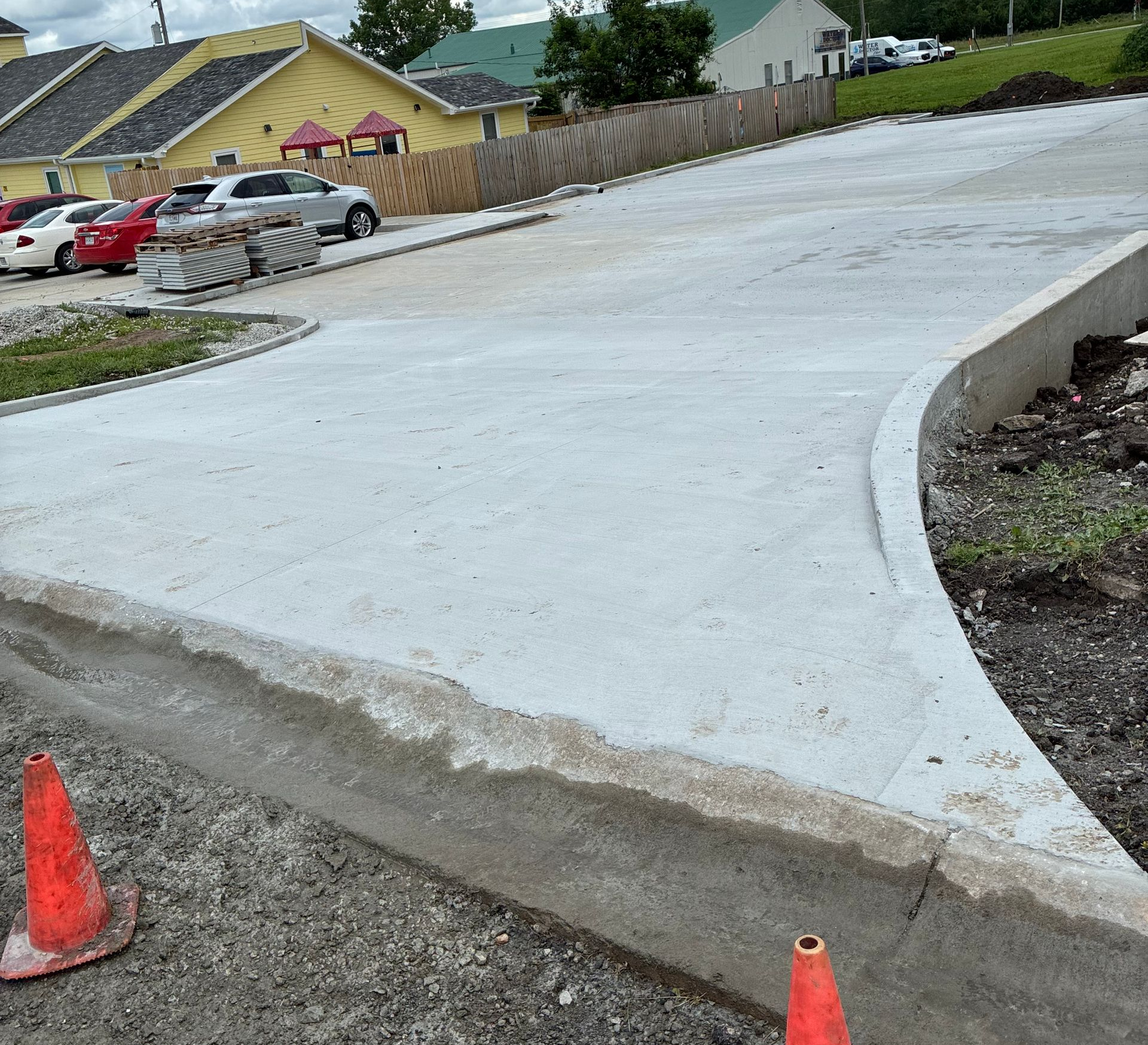 Curved white concrete path under construction beside a parking lot, with orange cones and a dirt edge