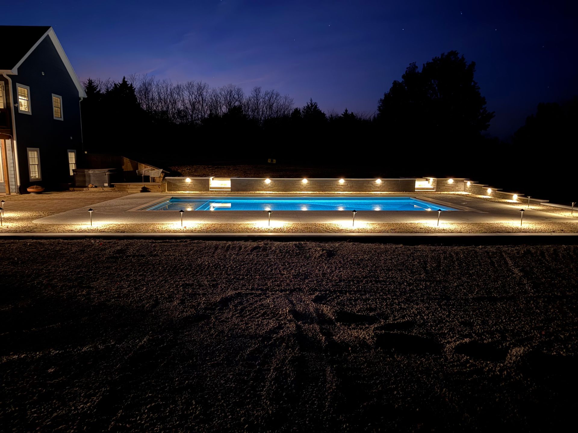 Nighttime backyard swimming pool with warm lights and a house on the left under a dark sky