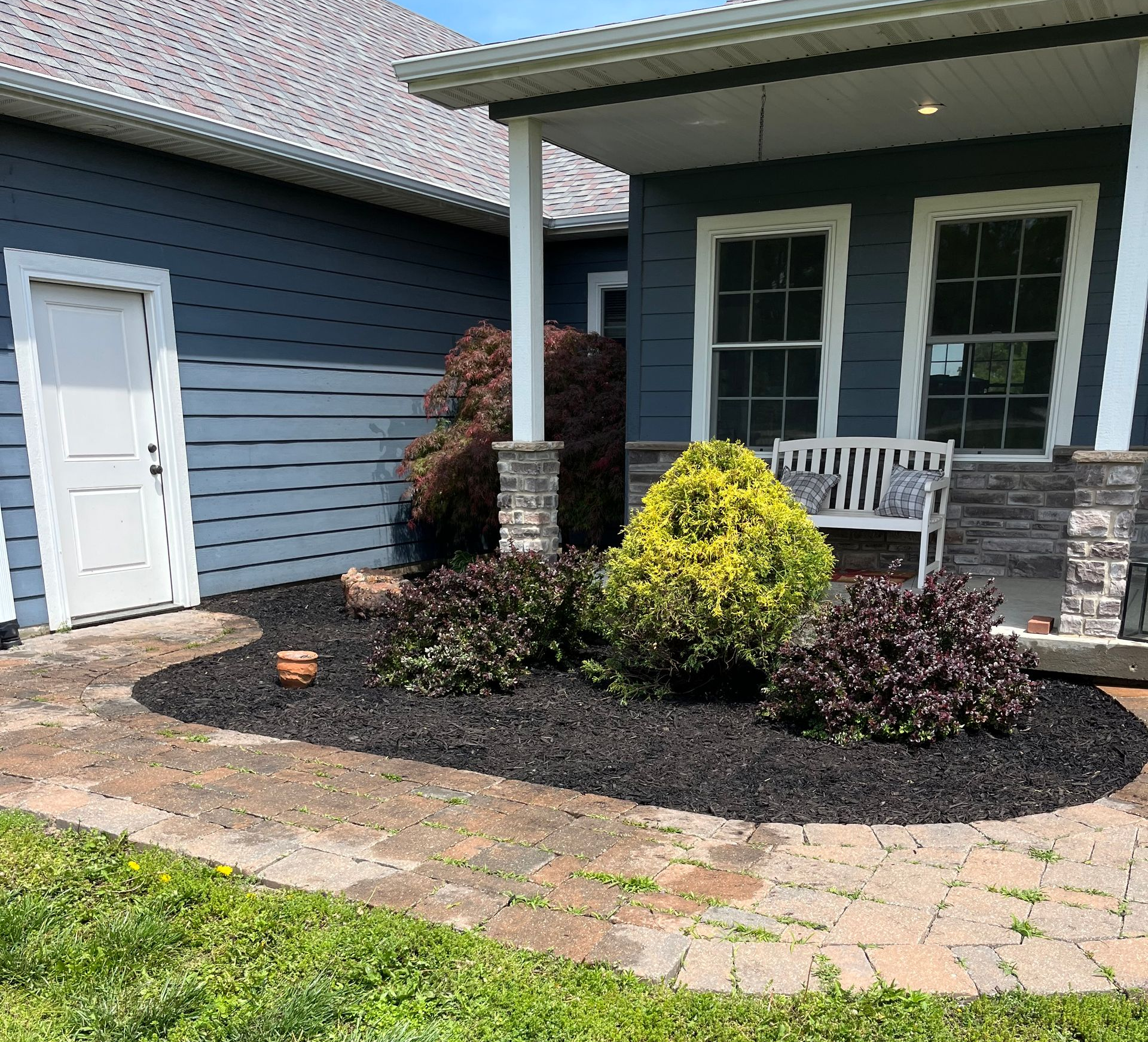 Front yard landscaping with mulch beds, green shrubs, and a house entry with gray siding and stone porch.