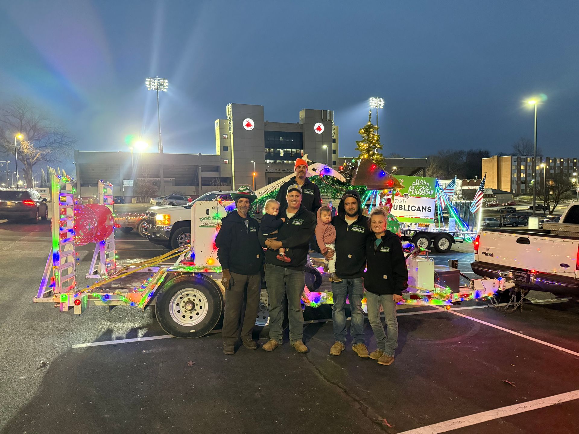 People posing beside a decorated parade float with colorful lights at night in a parking lot