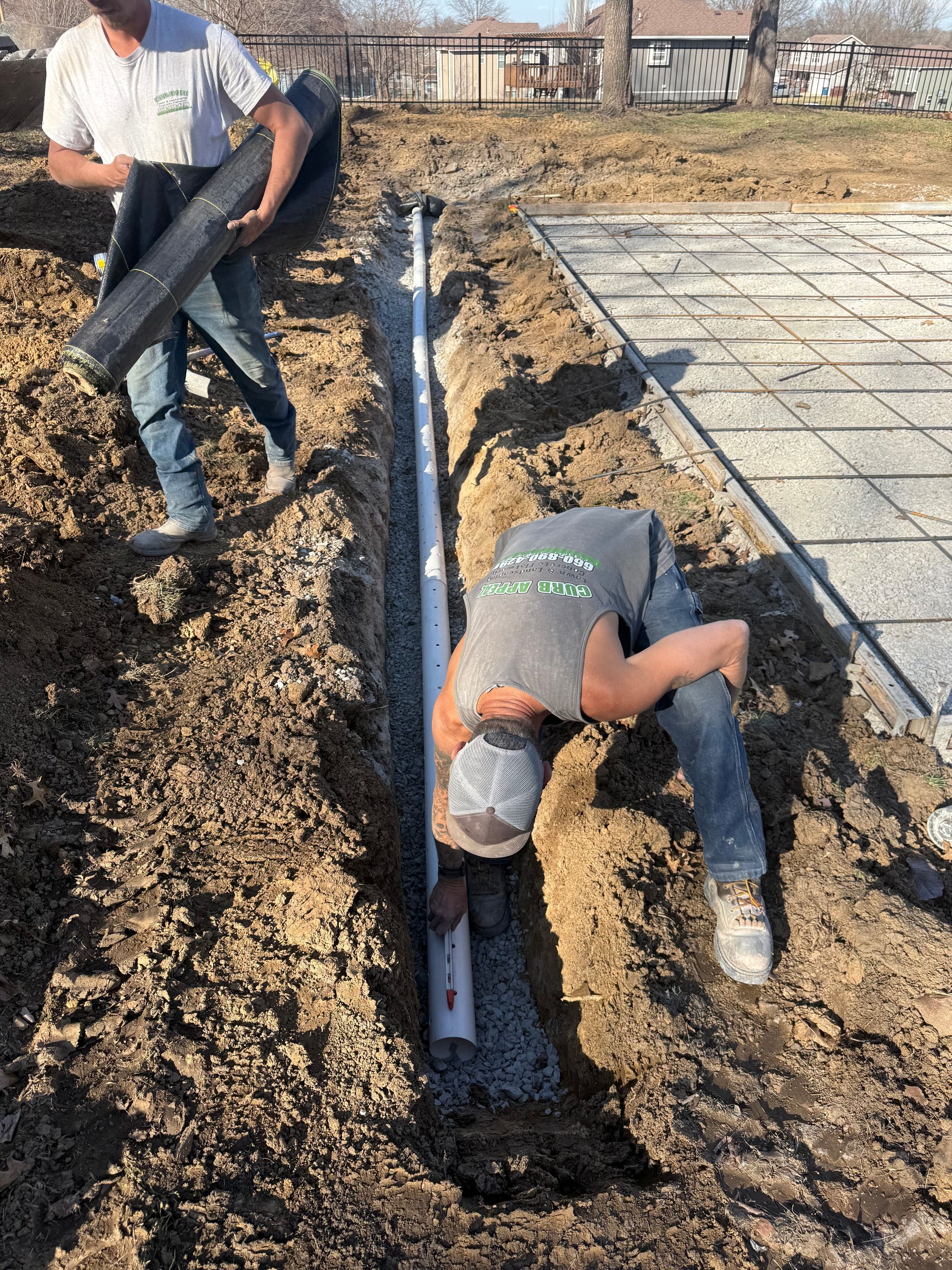 Two workers install a black drainage pipe in a muddy trench beside a concrete slab.