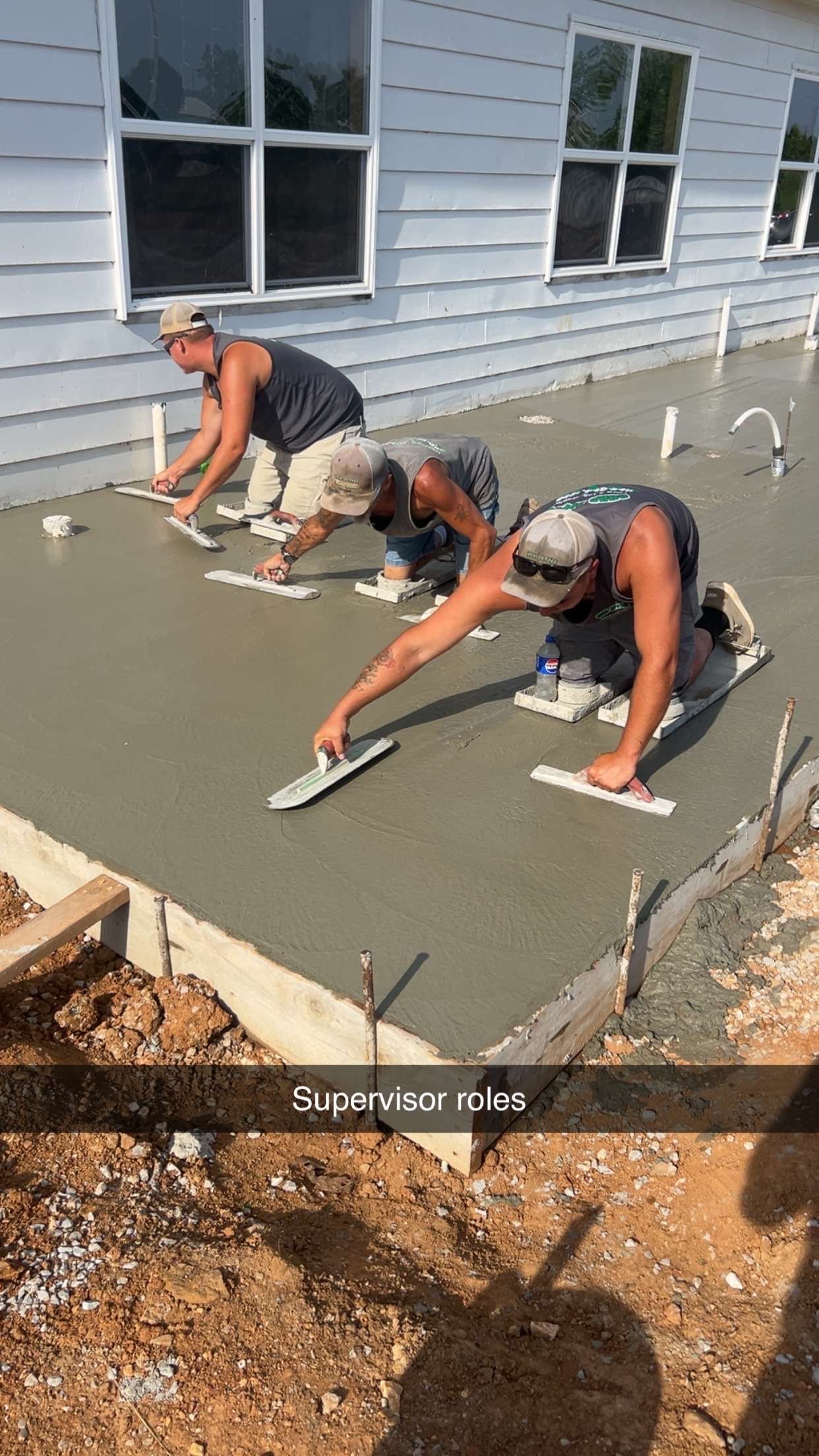 Two workers smoothing fresh concrete slab beside a house under construction.