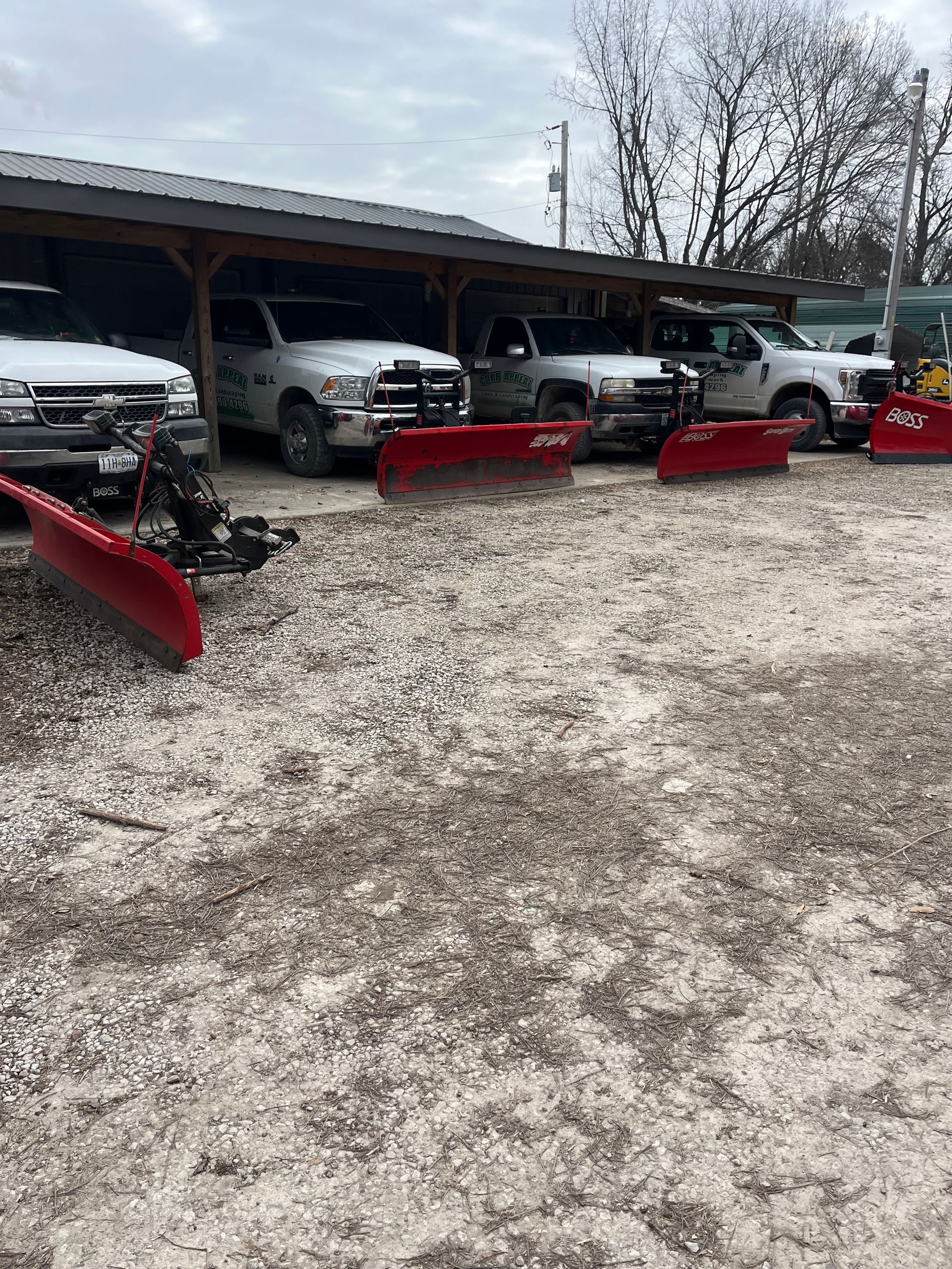 Red snowplows lined up under a shed beside white pickup trucks on a gravel lot.