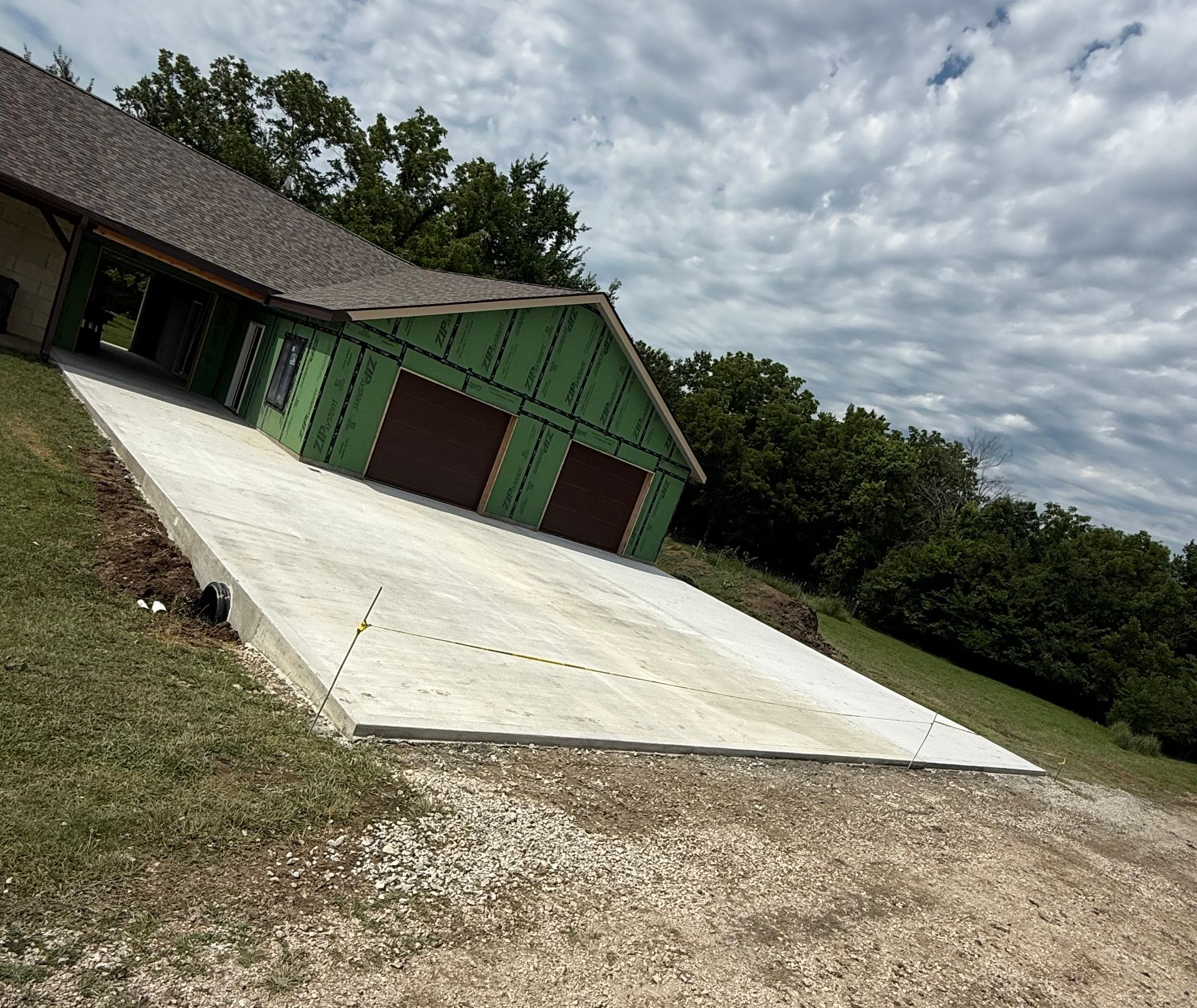 Slanted concrete driveway leading to a green garage with red doors beside a gravel yard under cloudy sky