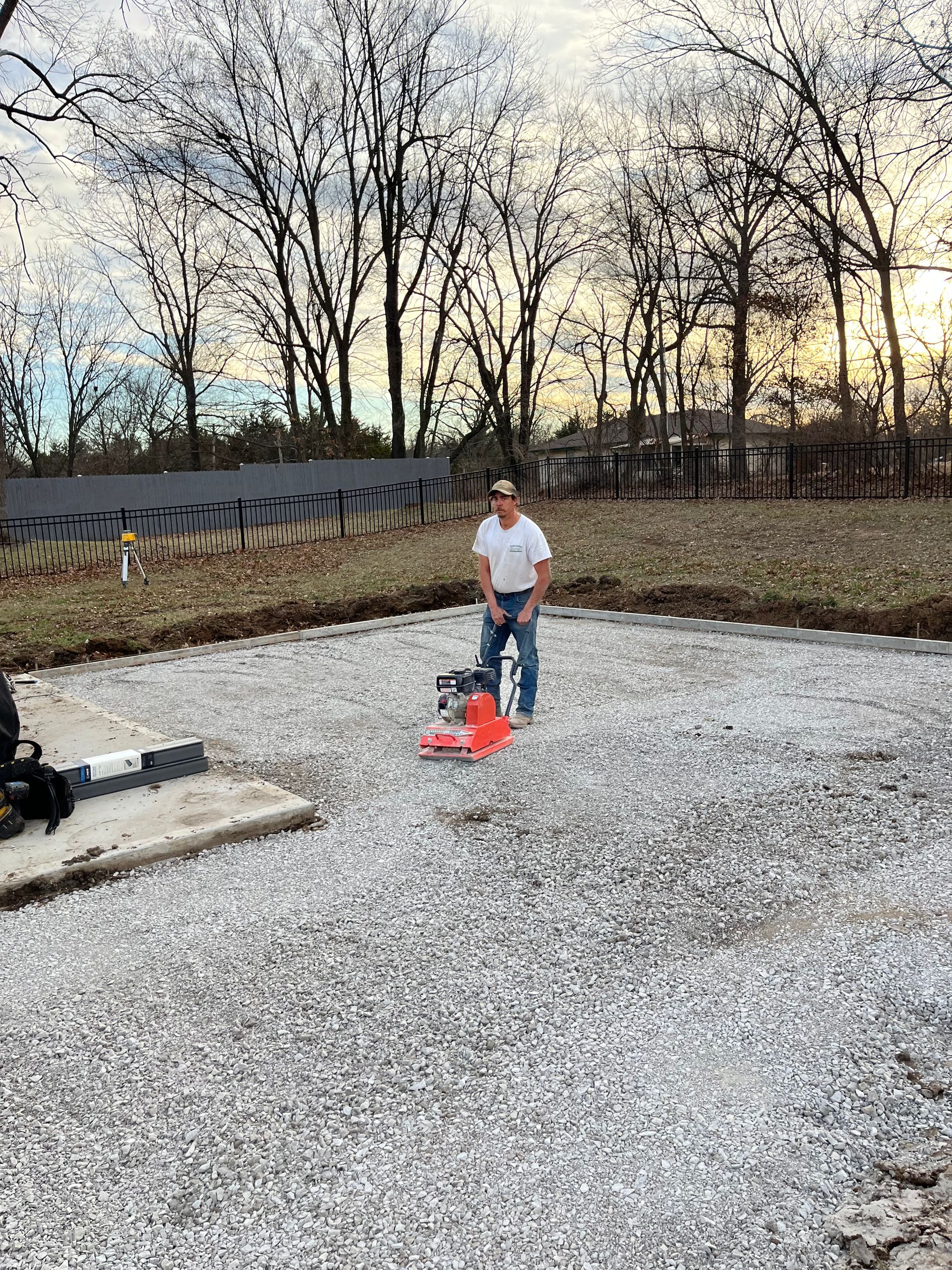 Person standing on gravel near a red lawn tool in an open yard with leafless trees at sunset