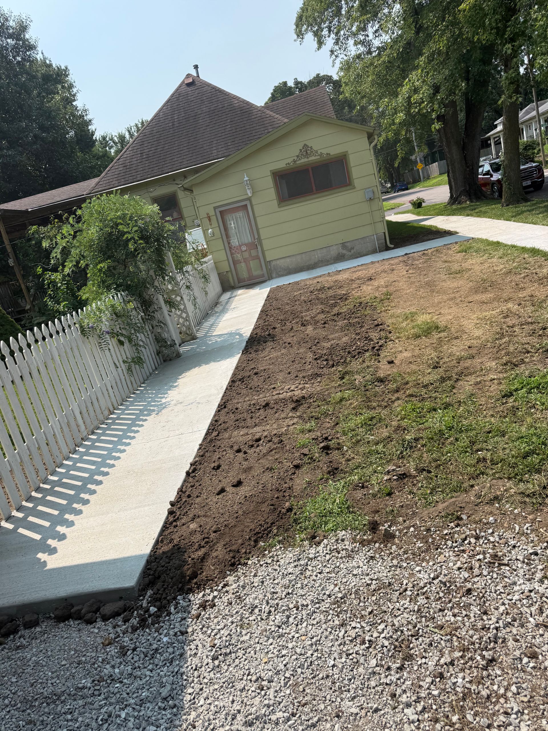 Tilted view of a small house with a red roof, white fence, gravel path, and a dry front yard under trees