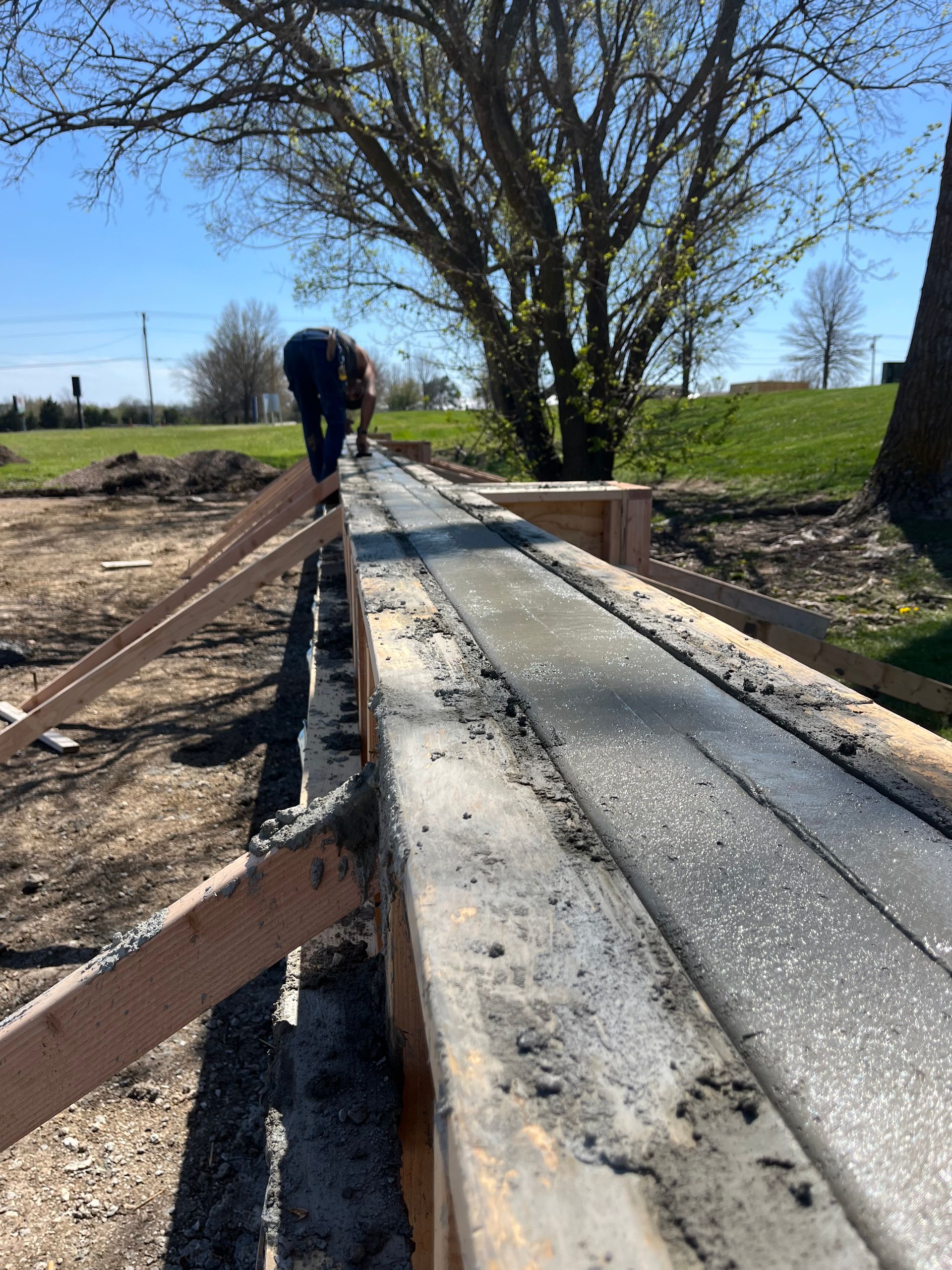 Person working on a narrow concrete bridge with wood rails in a rural field under a blue sky