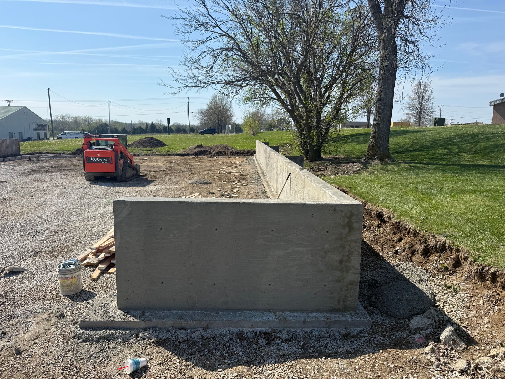 Concrete foundation wall at a gravel construction site with a red skid steer and trees in the background