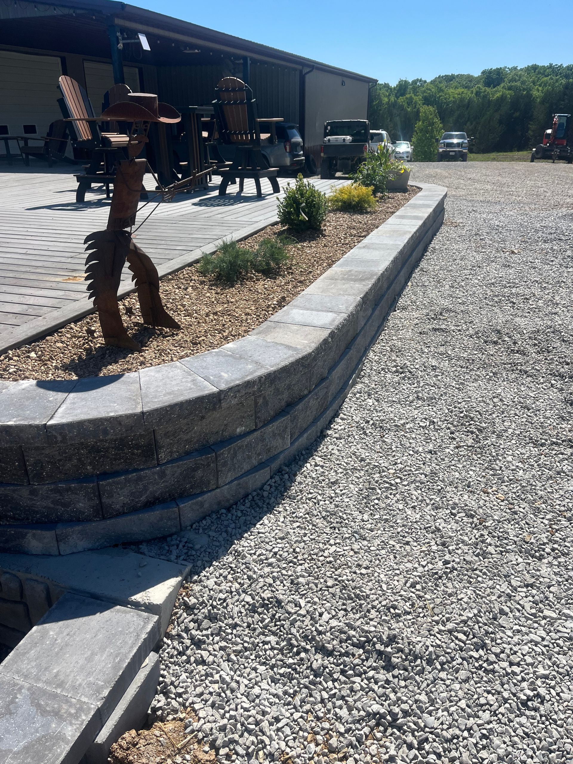 Curved stone planter with small shrubs beside a gravel patio and rustic outdoor seating area
