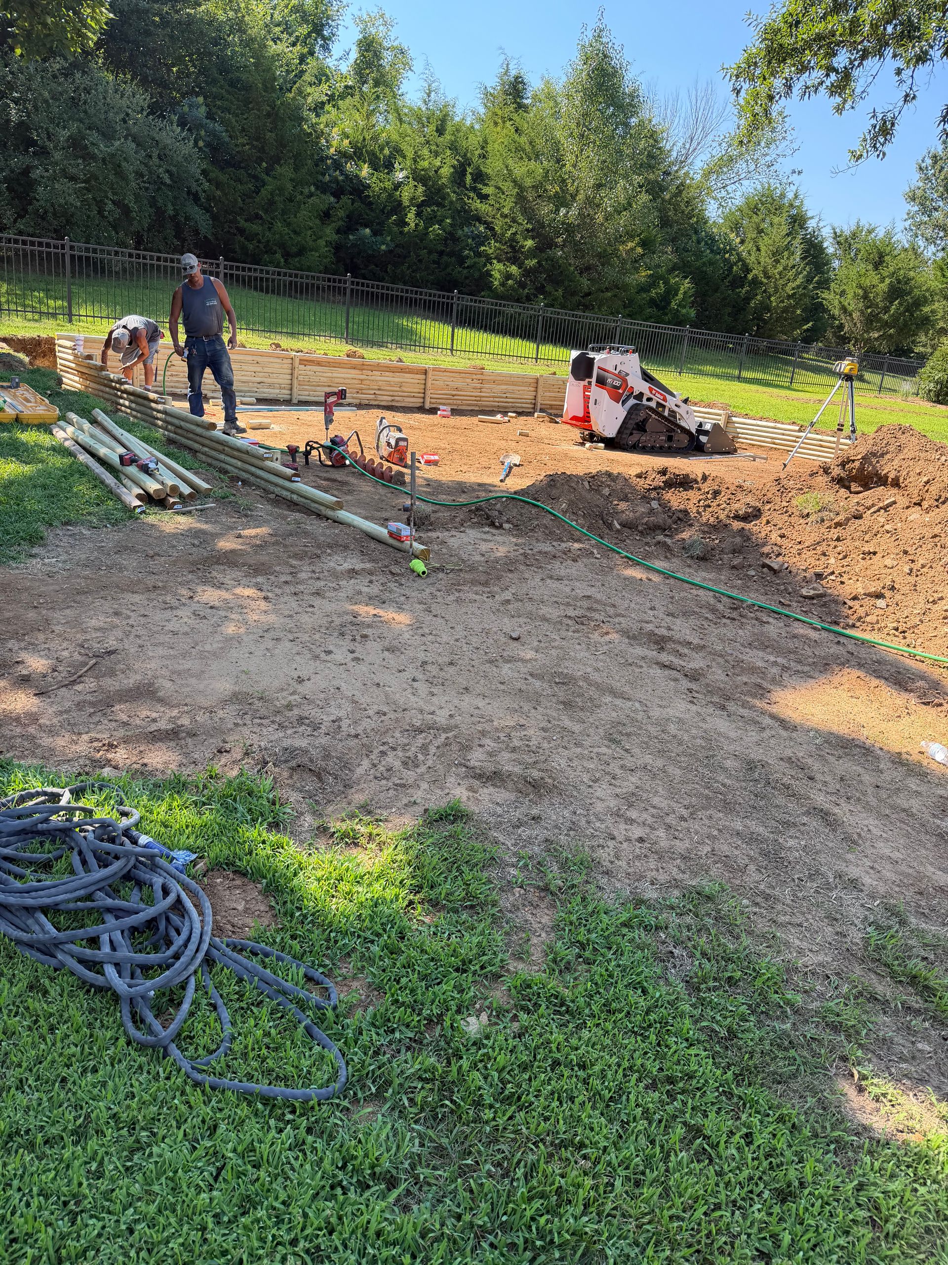 Workers at a muddy construction site with a small excavator, soil piles, and coiled hoses.