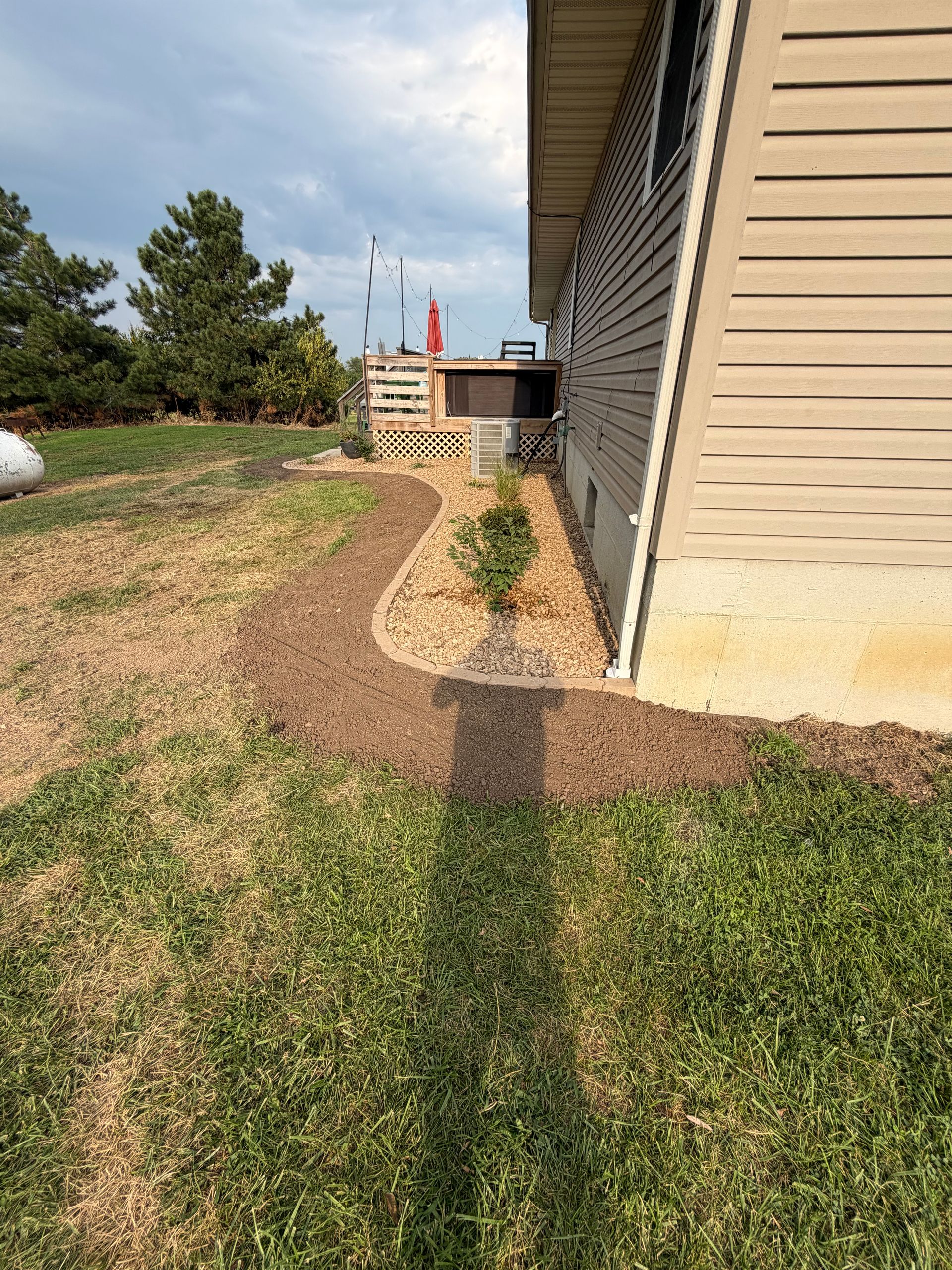 Sunlit backyard path beside a beige house, with grass, trees, and a small staircase in the distance