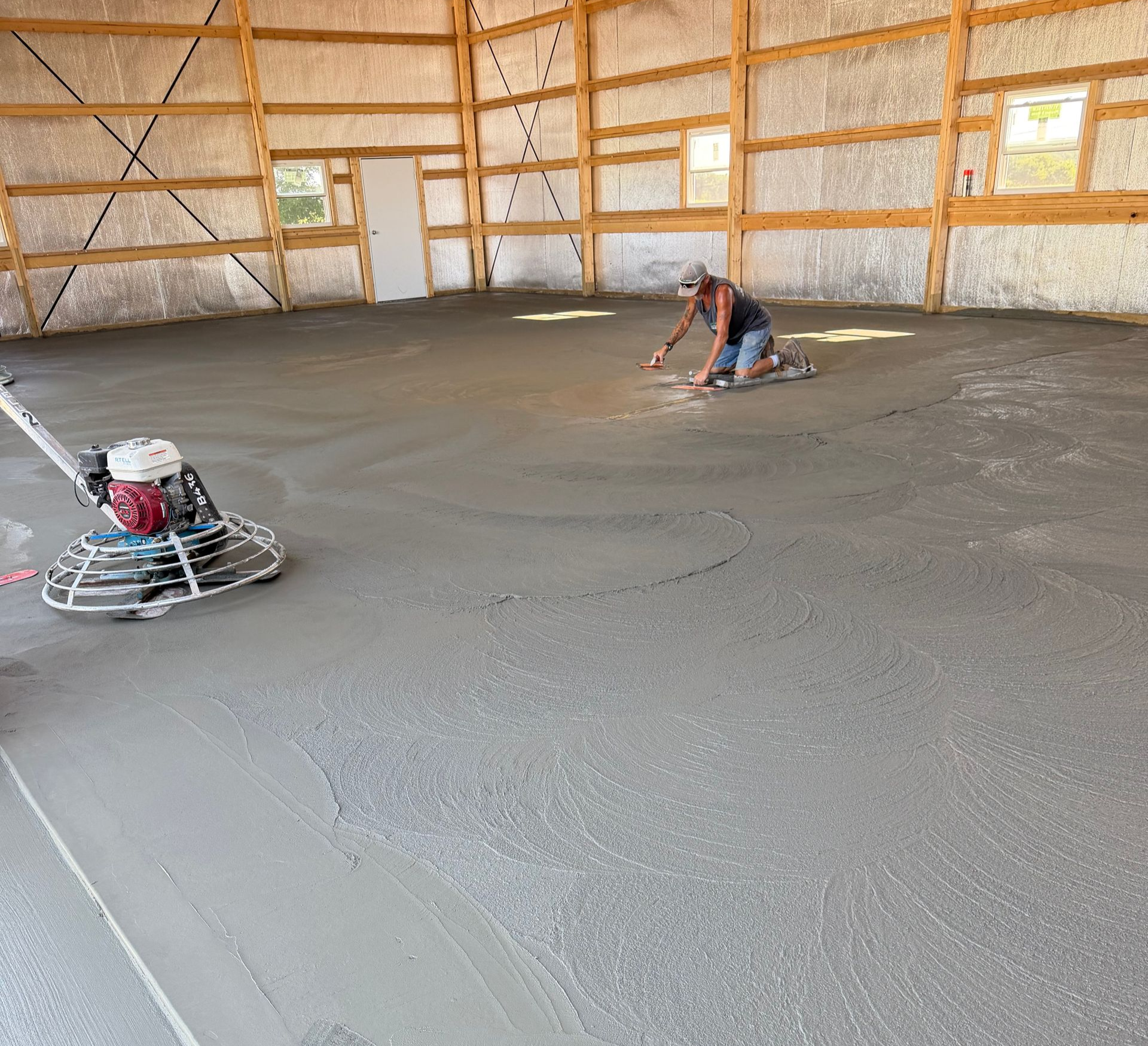 Workers smoothing fresh concrete floor inside a framed building interior.