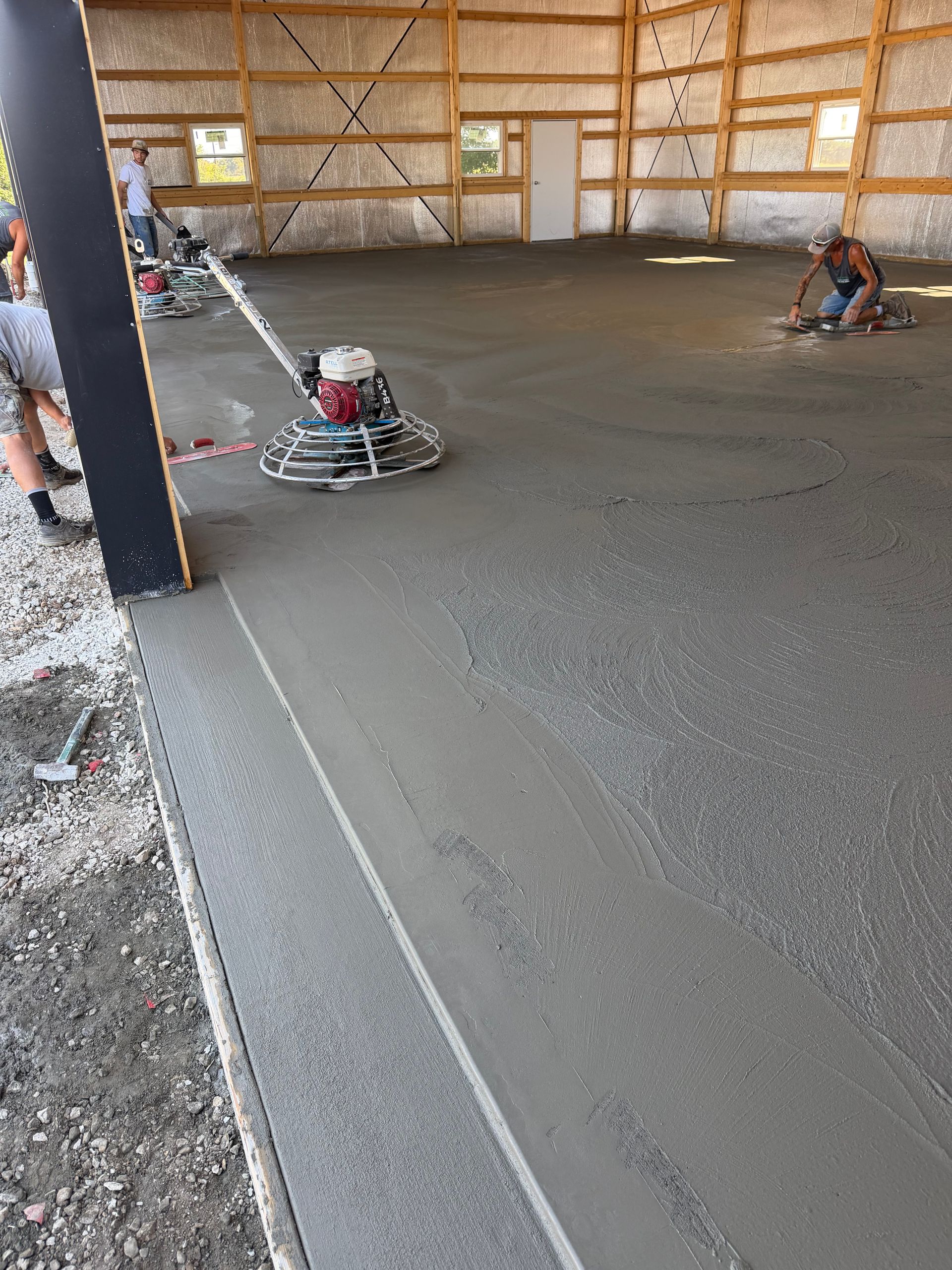 Workers smoothing fresh concrete floor inside a large wooden-framed building under construction