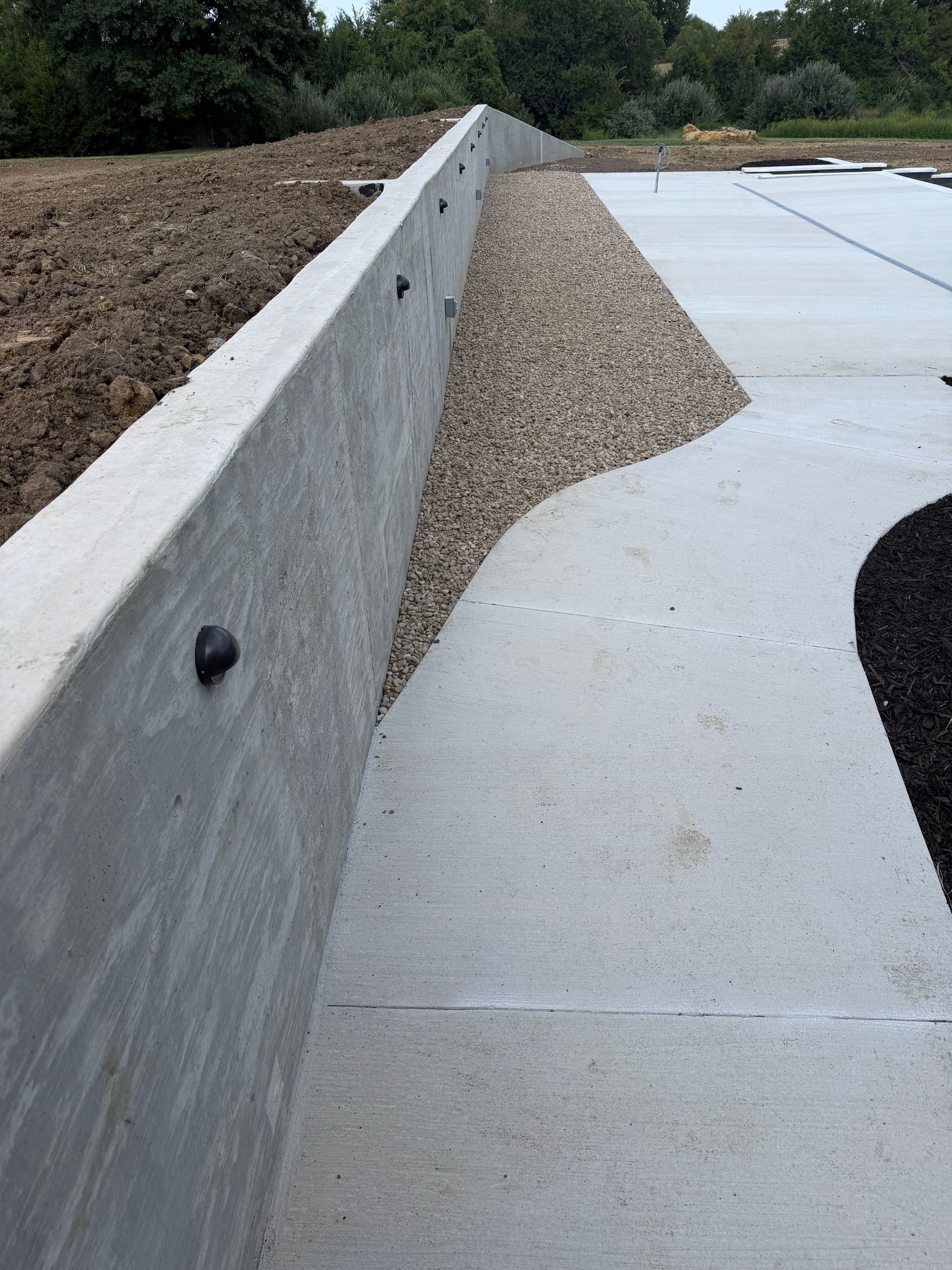 Concrete retaining wall with gravel spillover onto a paved surface beside landscaped ground.