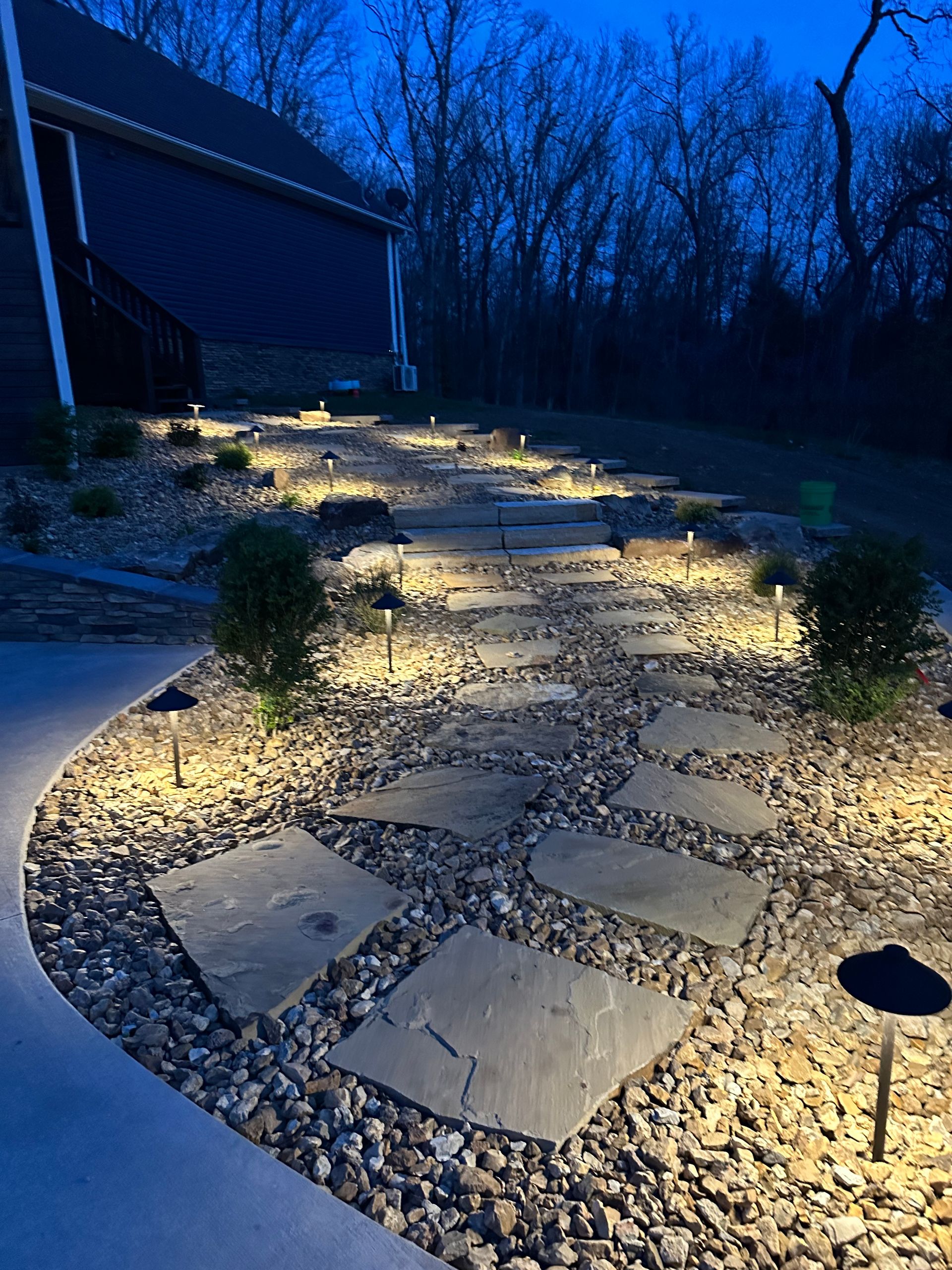 Illuminated stone path winding through gravel in a yard at dusk, with house on the left and trees in back