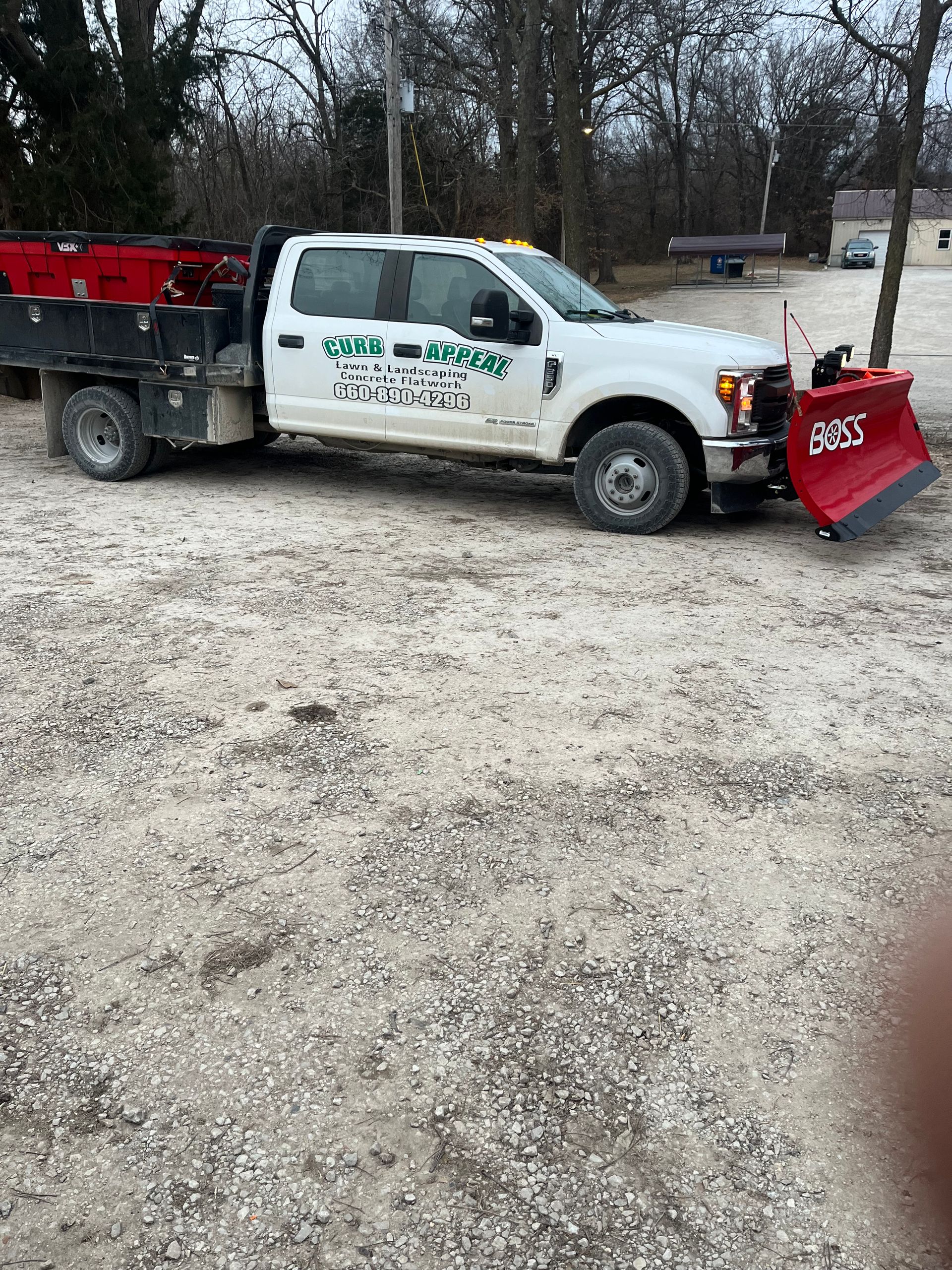 White utility truck with red snowplow parked on a gravel lot beside a wooded area
