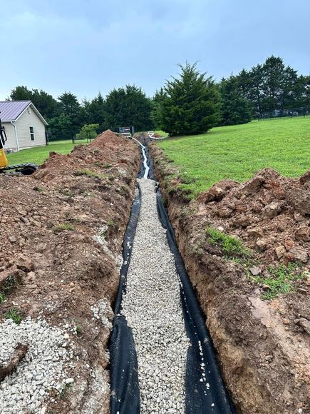 Gravel drainage trench through a grassy field beside a house, with fresh earth piled along both sides