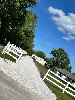 White fence along a gravel path in a sunny rural yard with green trees and blue sky